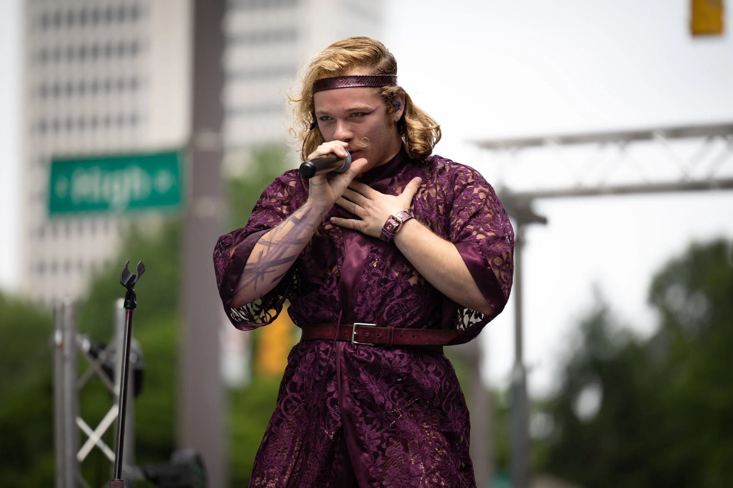 A person with long hair and tattoos, wearing a purple lace dress and headband, is singing into a microphone with one hand on their chest during an outdoor performance.