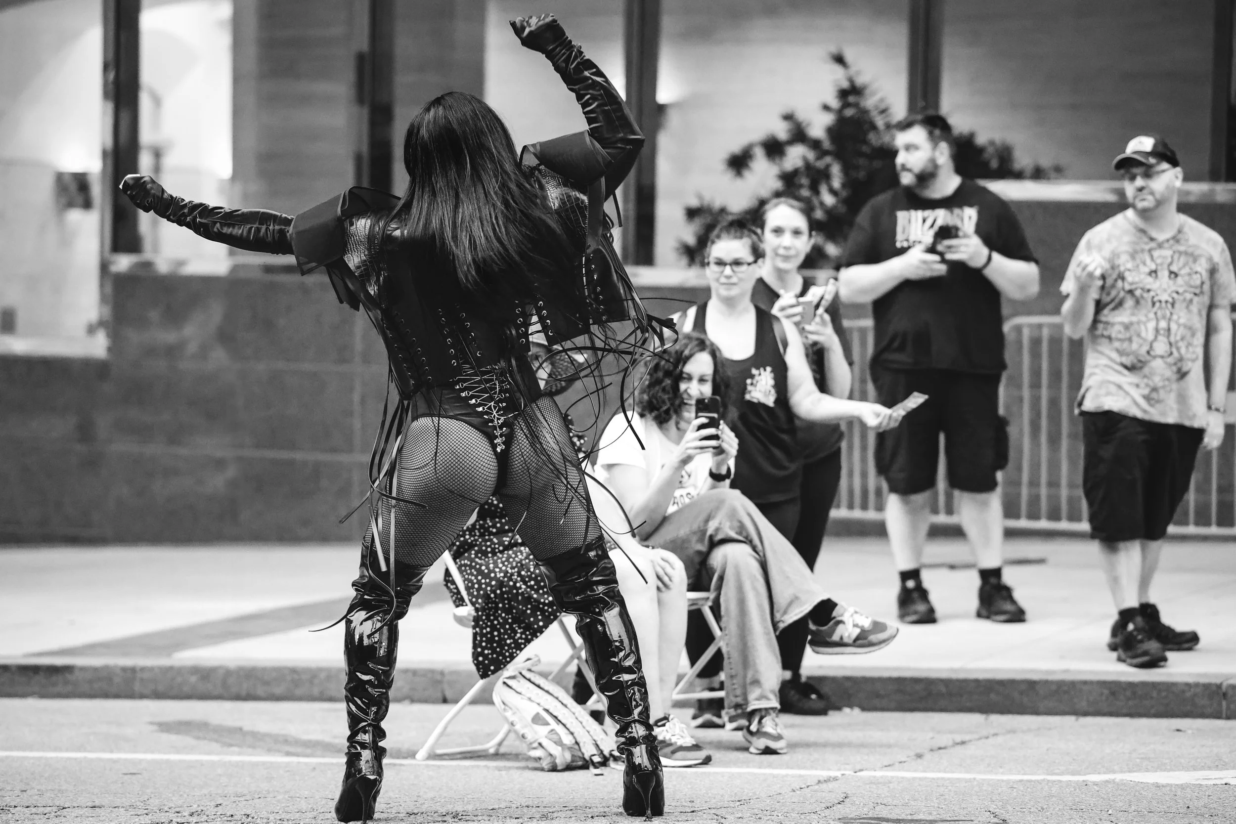 A performer in latex and fishnet clothing dancing or posing on the street with an audience taking photos and videos in the background.