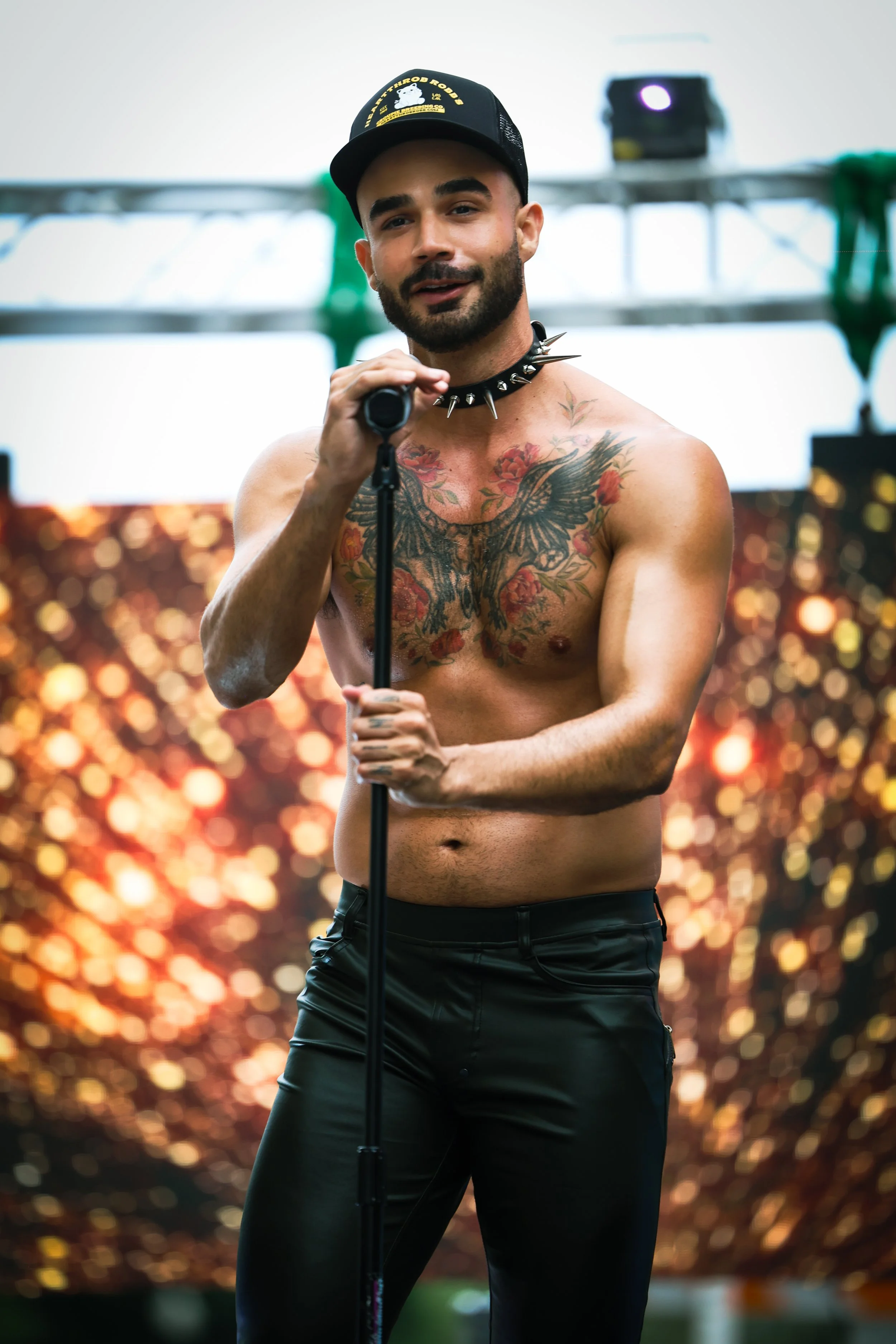 A shirtless man with tattoos, wearing a black cap and leather pants, holding a microphone on a stage with a colorful, blurred background.