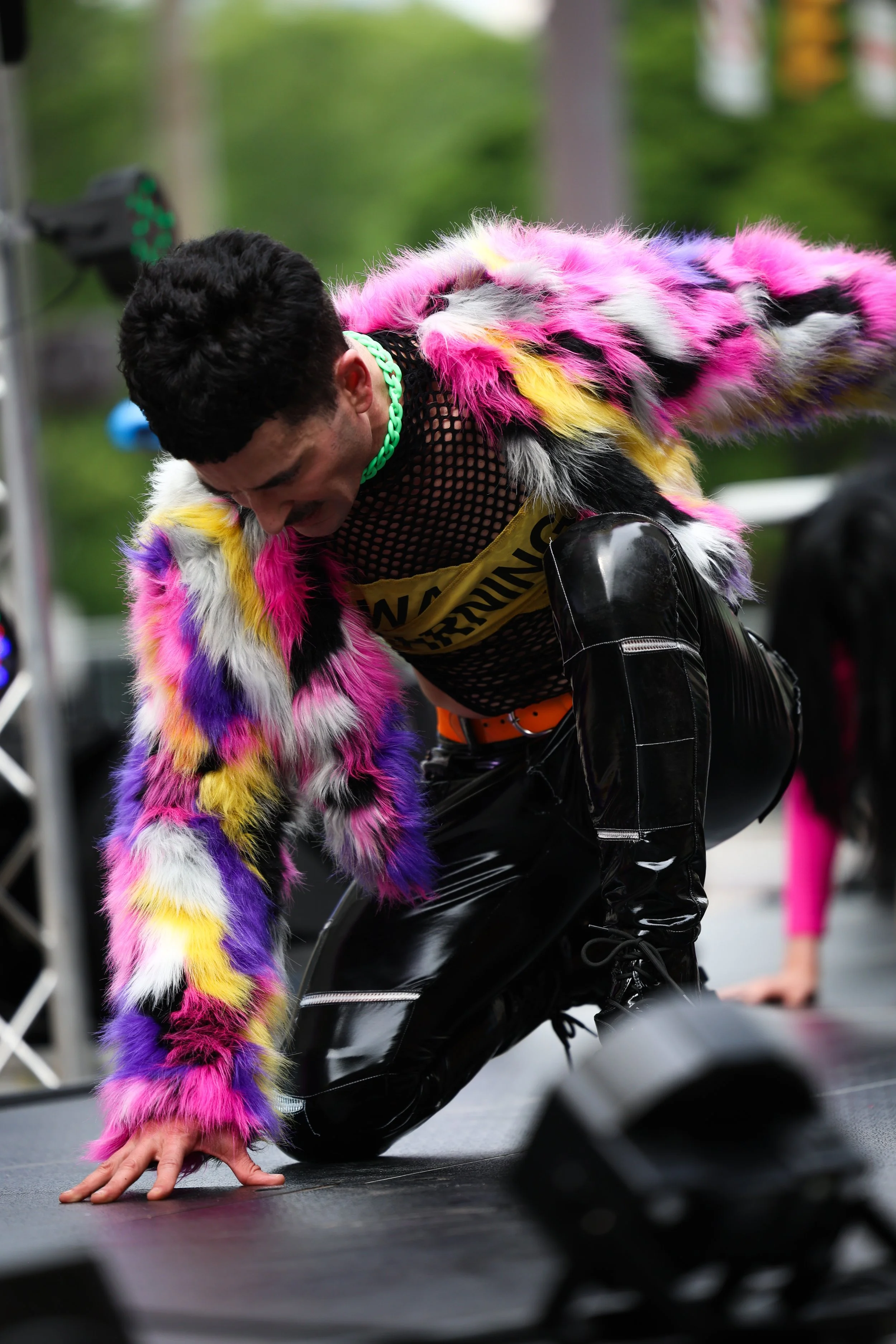Person wearing a colorful, fuzzy faux fur coat, black fishnet top with a 'WARNING' sign, and shiny black pants, crawling on all fours on a stage during an outdoor event.