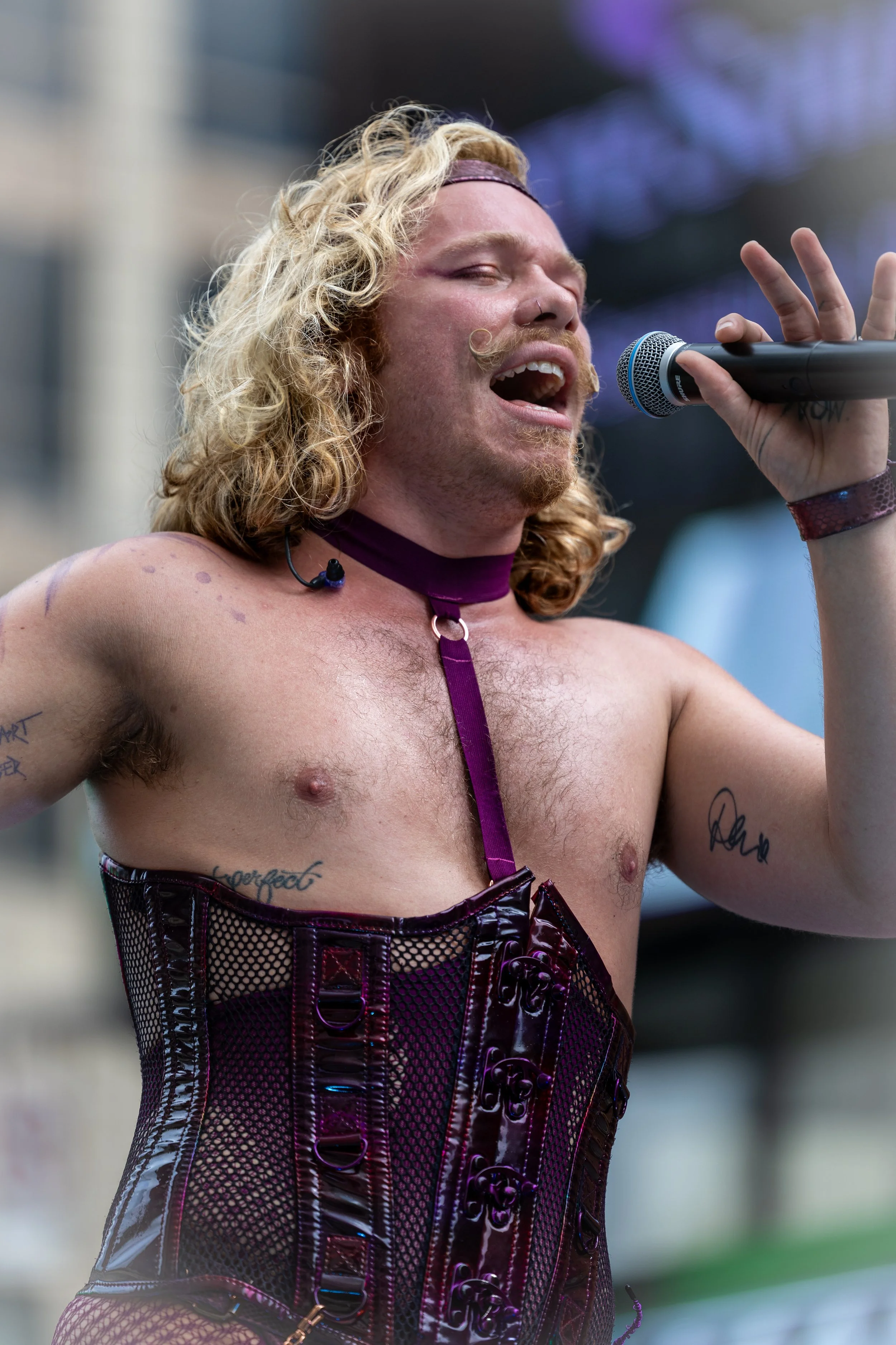 A person singing passionately during a performance, wearing a purple choker and corset, with tattoos on their arms.