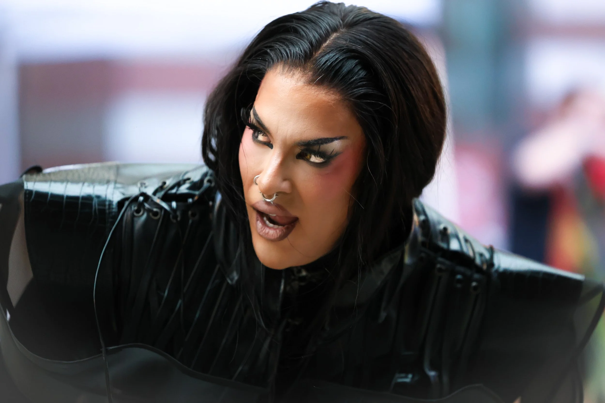 A woman with dark, styled hair, makeup, and multiple facial piercings, wearing a black outfit with shiny details, is looking to the side with an intense expression.