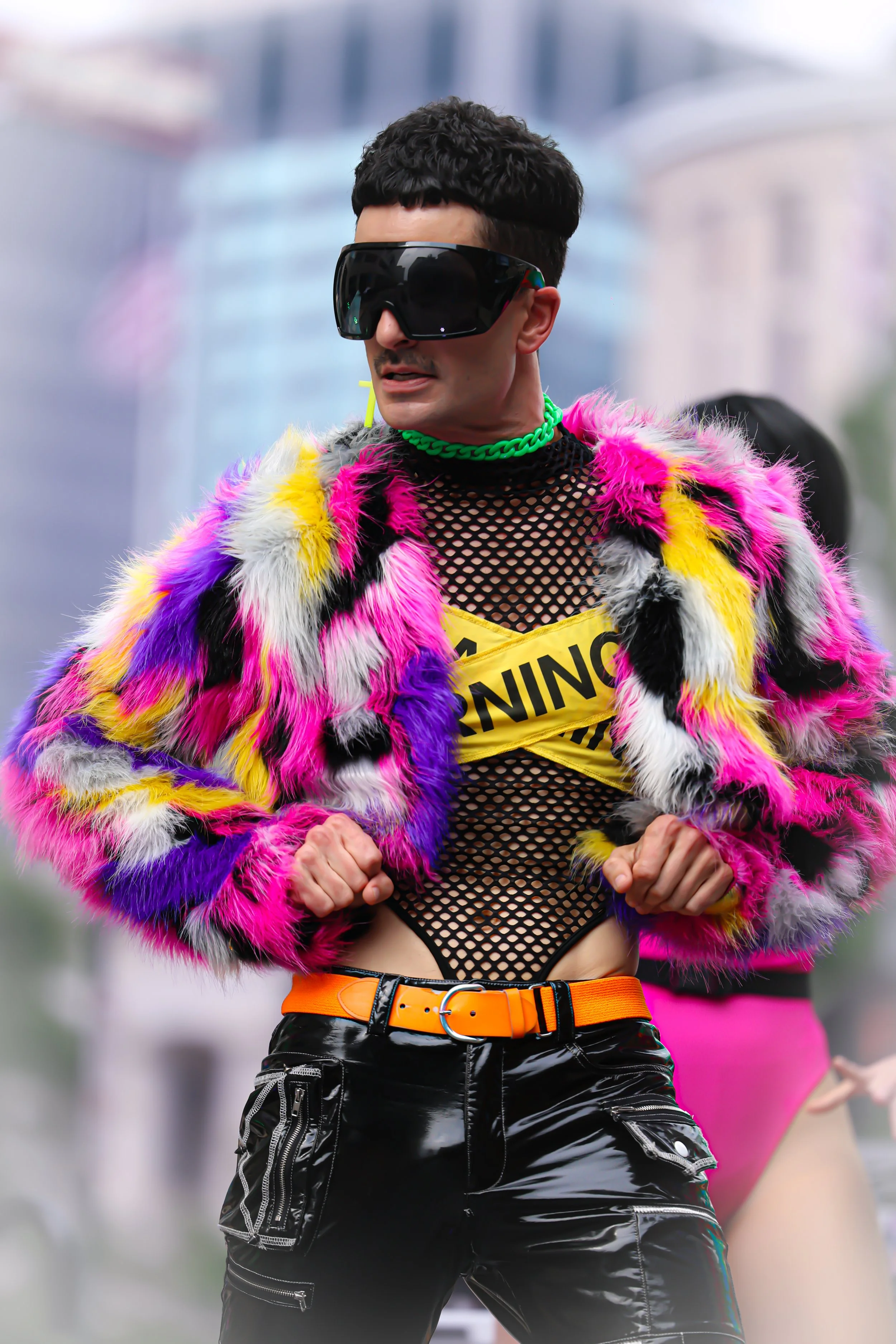 Person in a colorful, furry jacket and black fishnet top with a yellow 'Caution' sign, wearing futuristic sunglasses and posing outdoors.