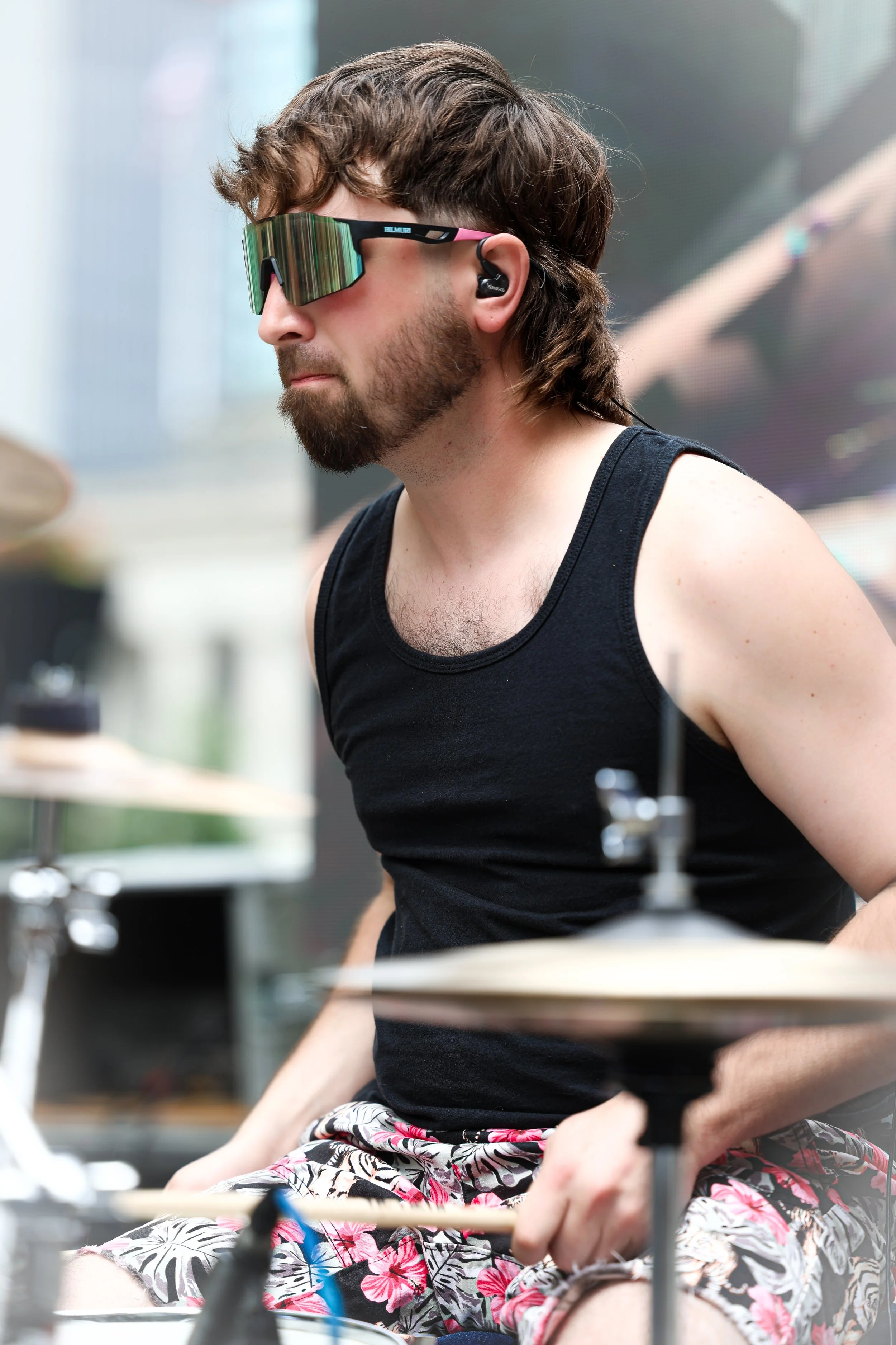 A man with a beard and long brown hair playing drums, wearing sunglasses, a black tank top, and shorts, with a blurred outdoor background.