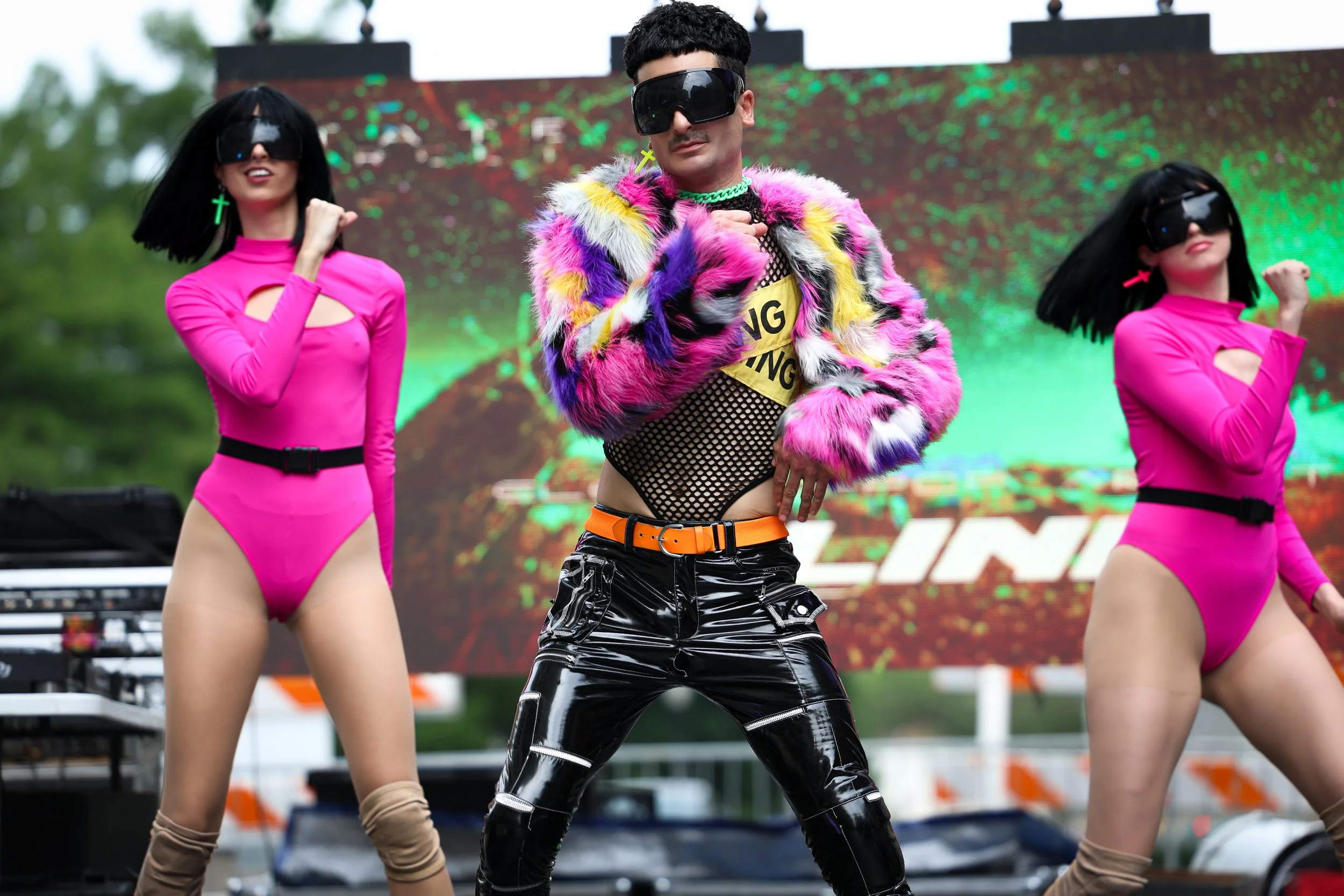 Performer in colorful fur coat and black vinyl pants dancing on stage with two backup dancers in pink bodysuits, all wearing black visors, in an outdoor setting with a digital background.