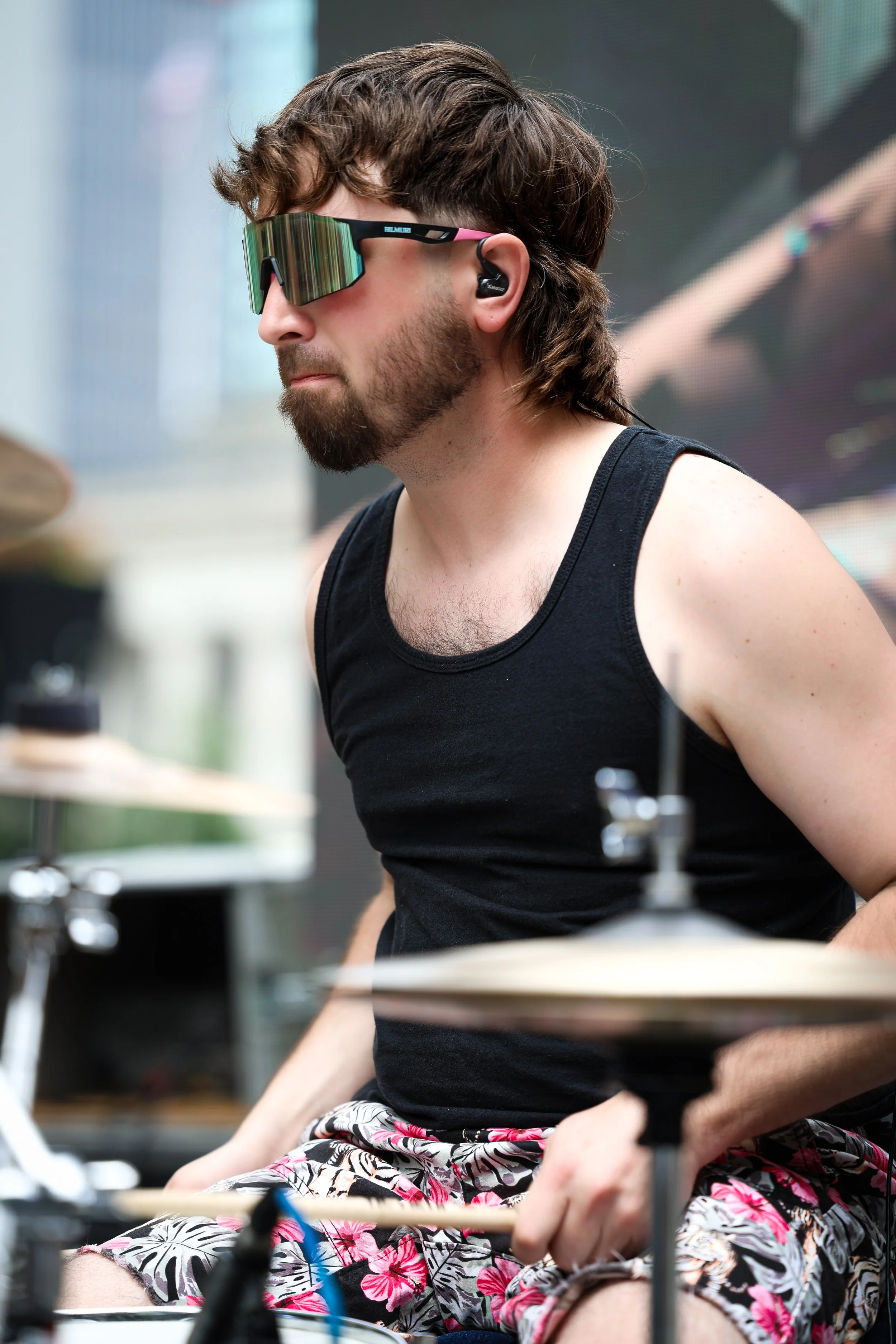 A man with long wavy brown hair, beard, wearing sunglasses, a black tank top, and floral shorts, appears to be playing drums.