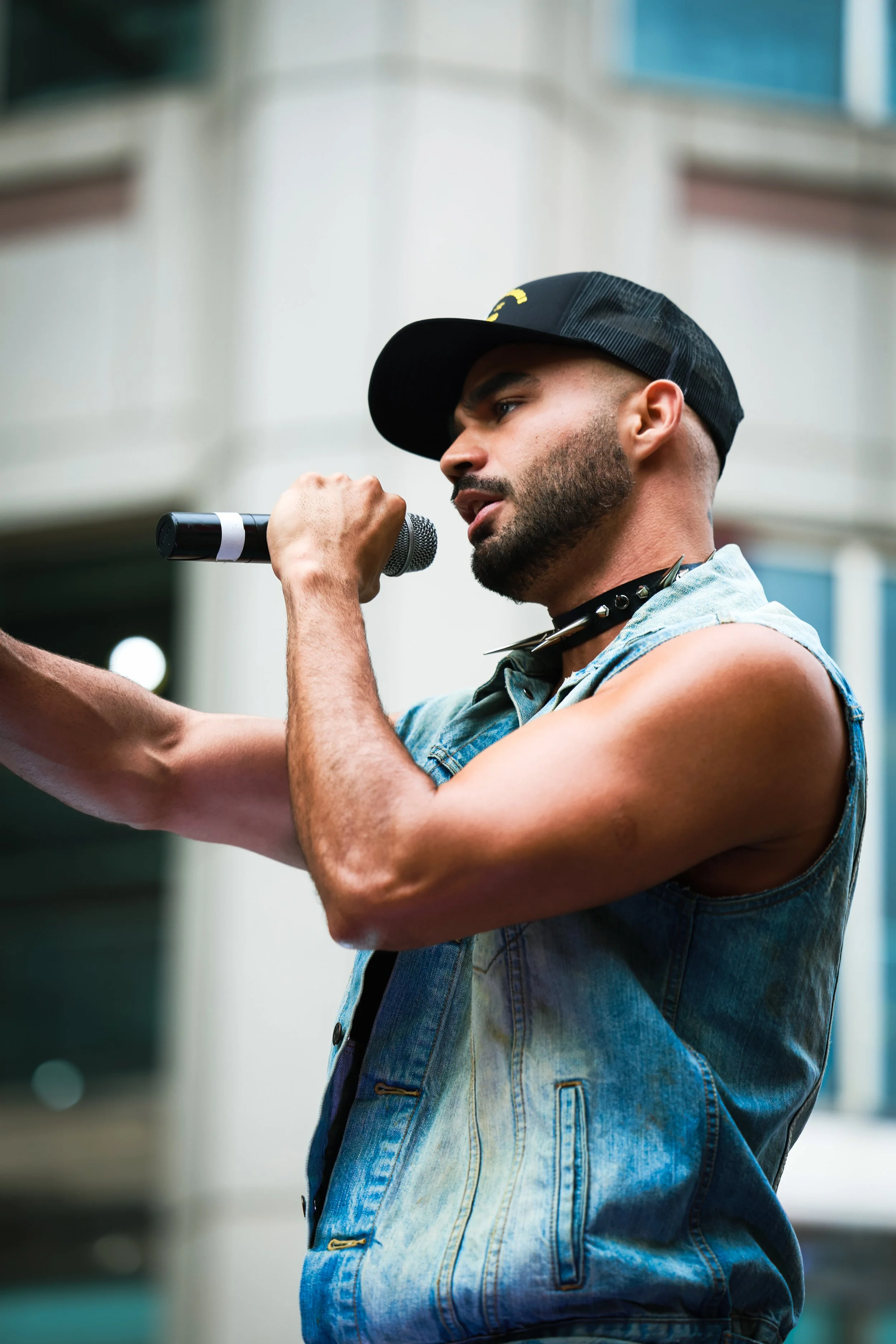 A young man with a beard, wearing a denim vest, a black cap, and a spiked choker, is speaking into a handheld microphone during a public performance or protest, with a city building in the background.
