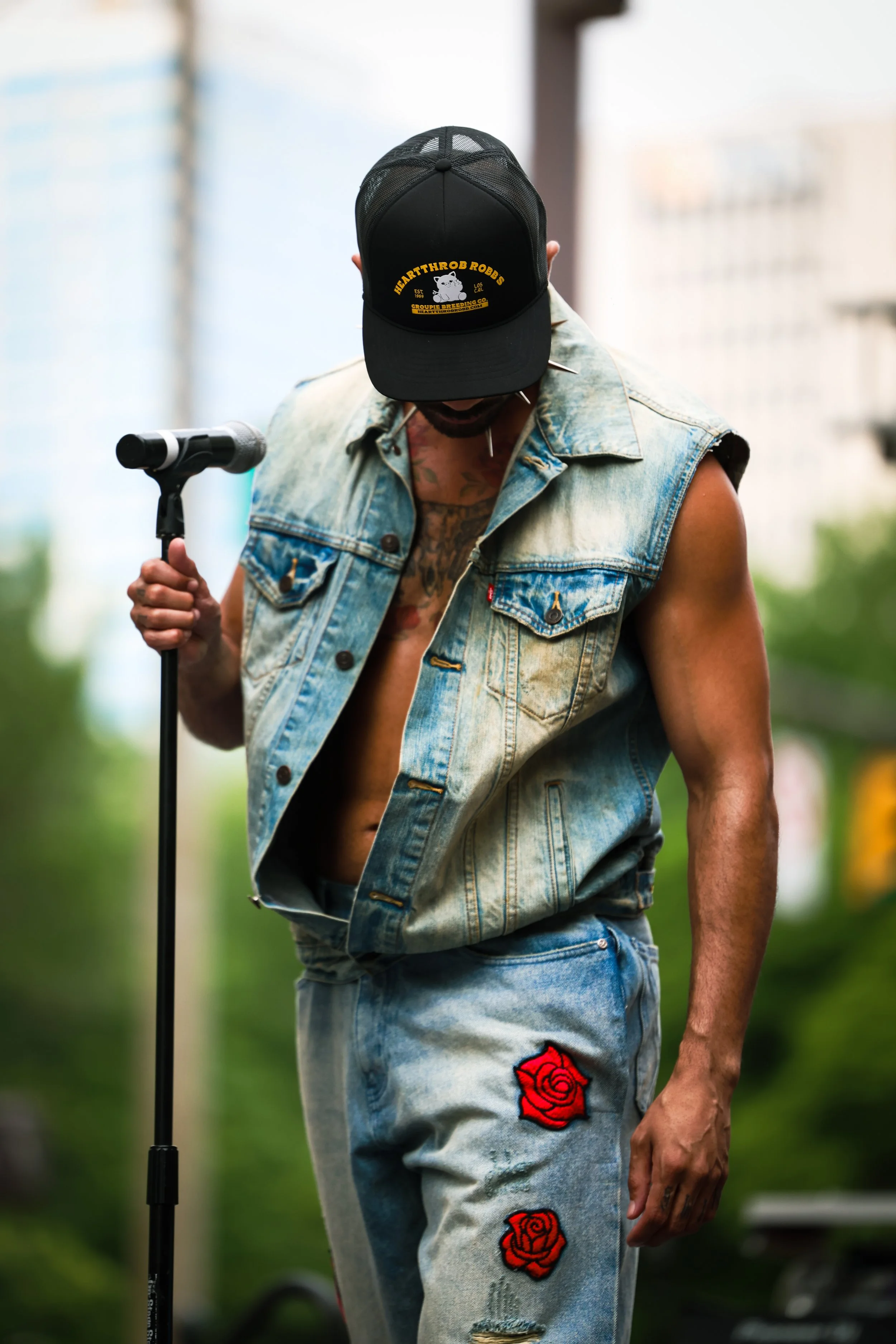 A model standing outdoors, wearing sleeveless denim vest and distressed jeans with rose patches. The model is holding a microphone in one hand and is wearing a black cap with yellow text.