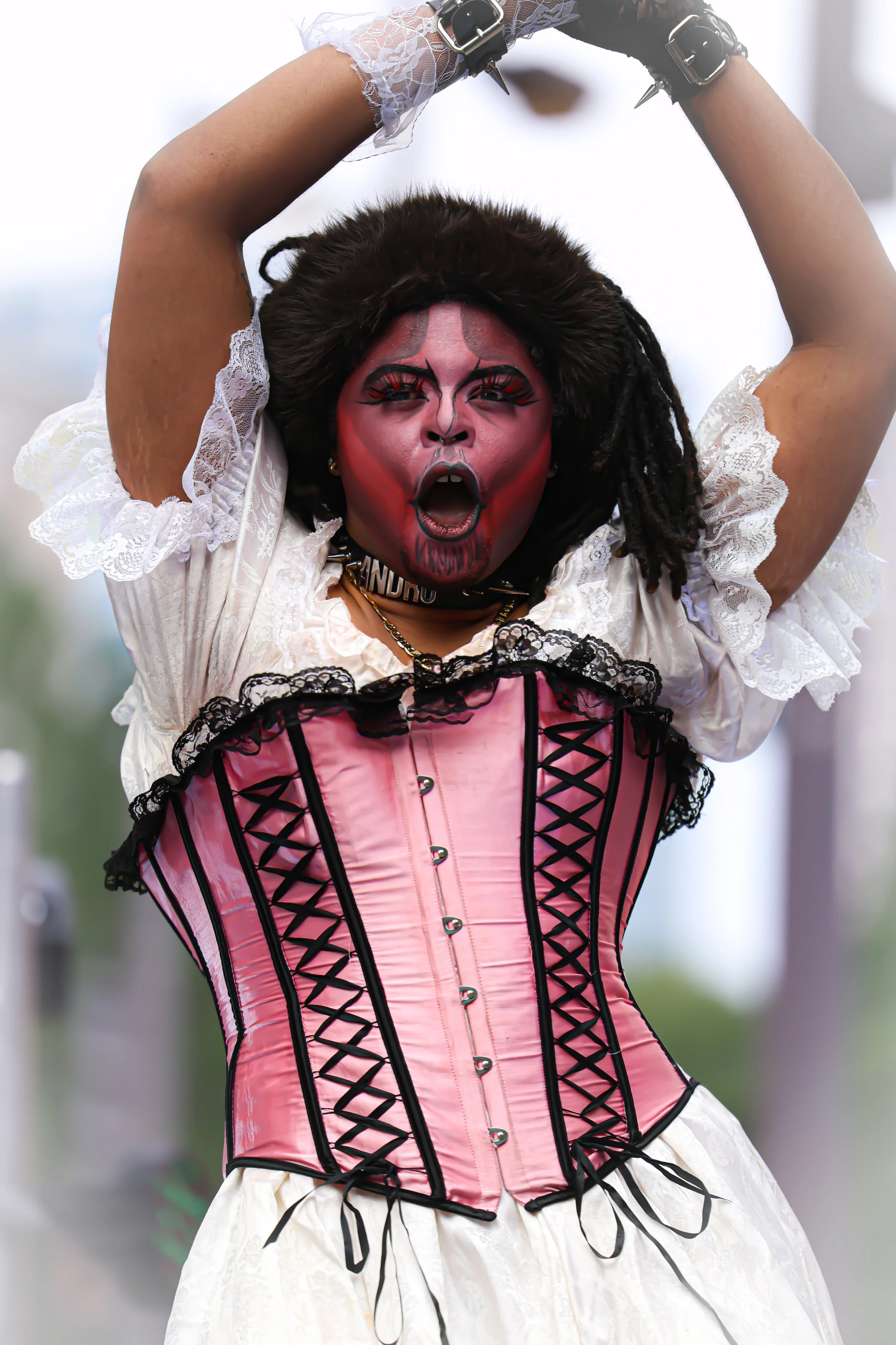 A person dressed in a Gothic-style costume with a pink corset, lace and ruffles, and a skull choker, with face makeup resembling a red demon or vampire, during a performance or event.