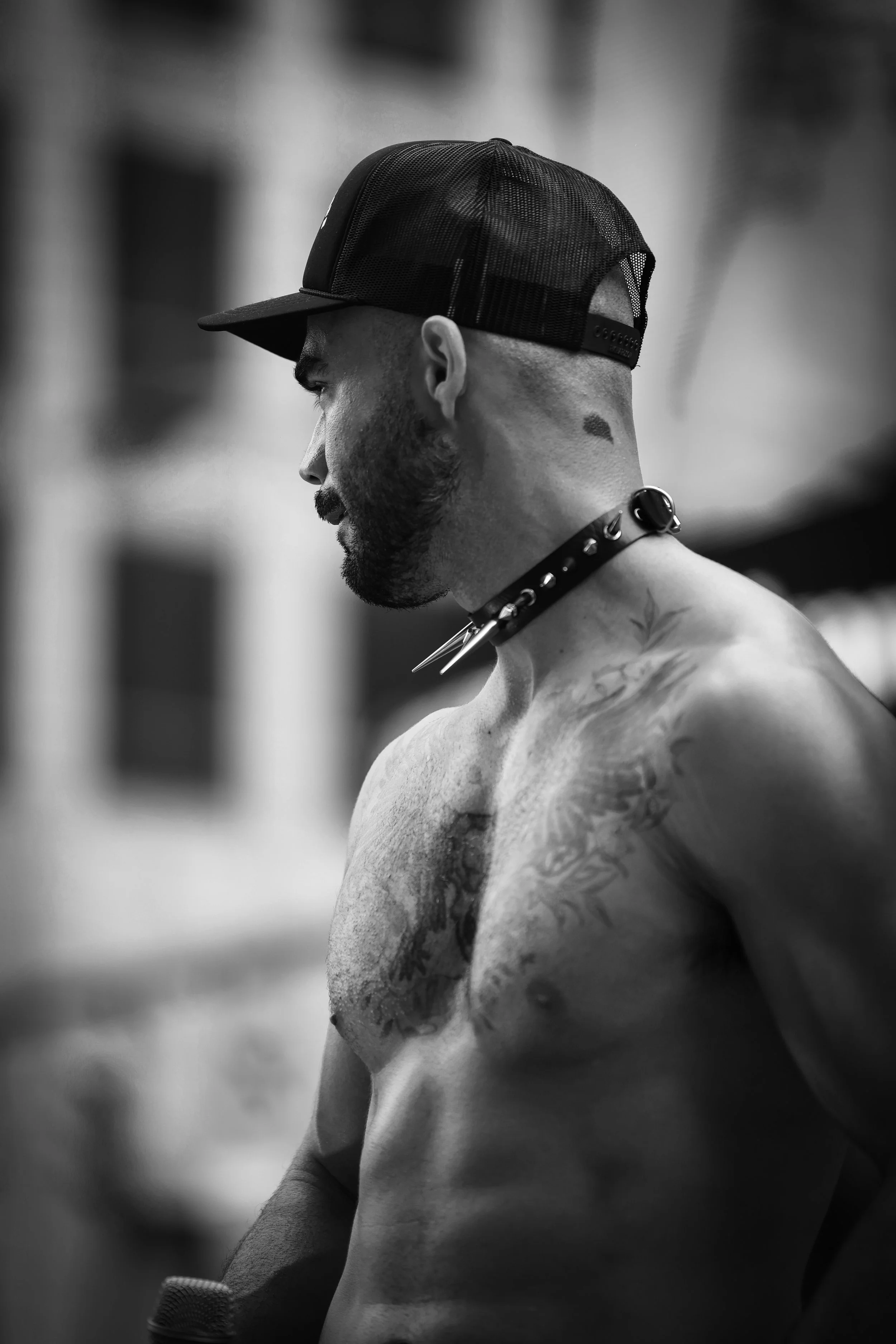 A shirtless man with tattoos on his chest and arms, wearing a baseball cap, a spiked collar, and a choker, standing in profile against a blurred urban background.