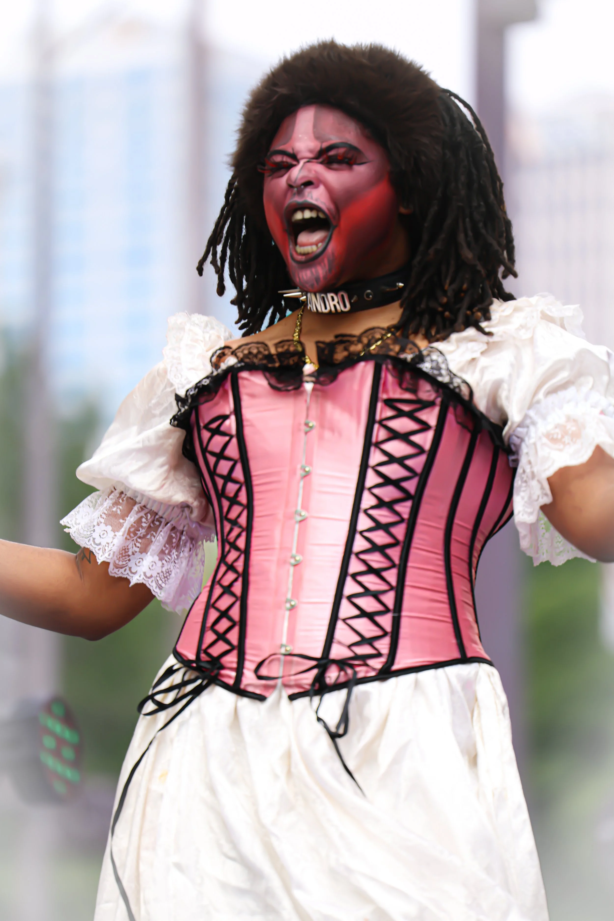 Person wearing a pink corset, white lace blouse, and white skirt, with dramatic makeup and a fierce expression, performing on stage.