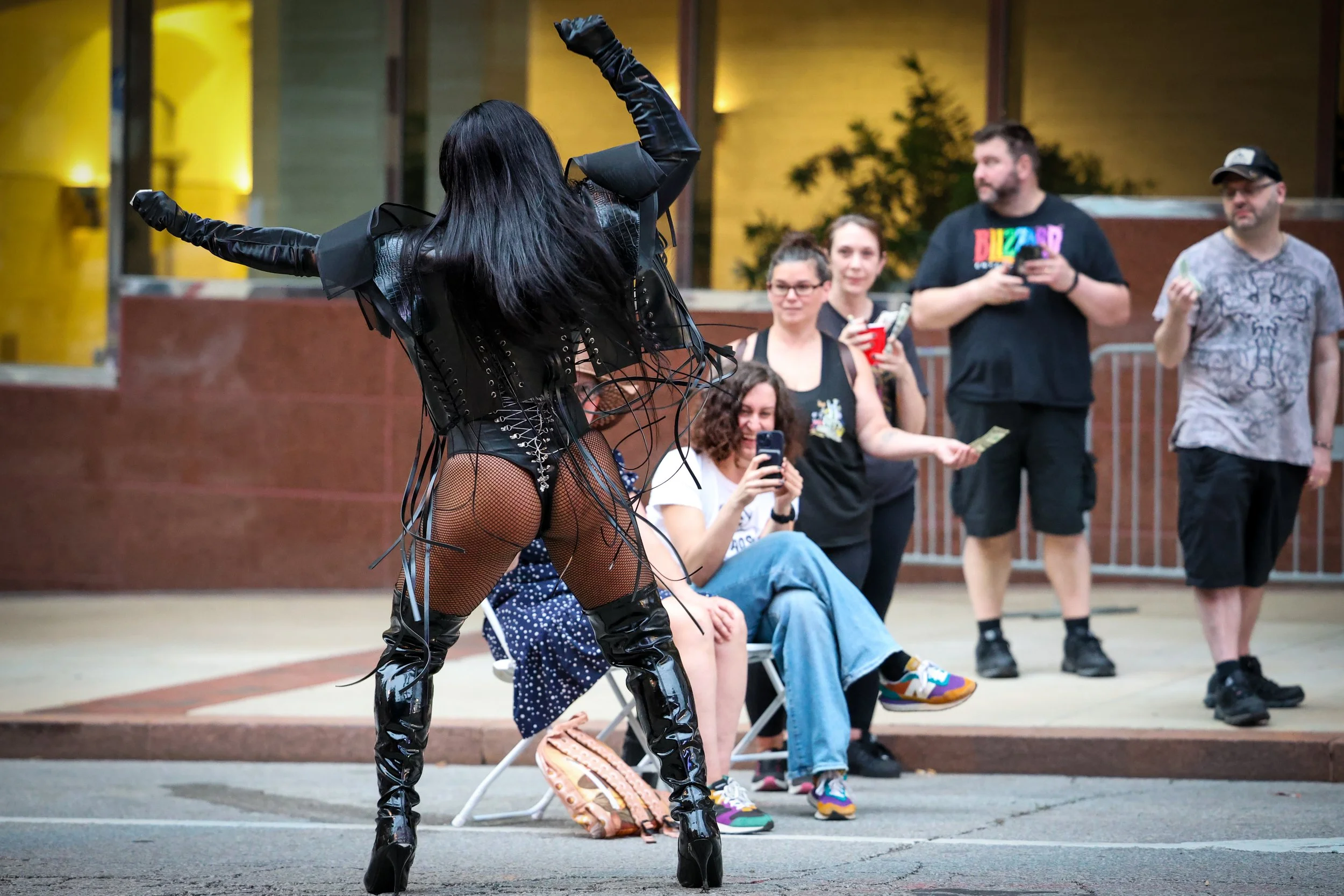 A performer dressed in black leather and fishnet with thigh-high boots dances in front of an audience. The audience members are watching and taking pictures, some sitting and some standing, at an outdoor event during evening.