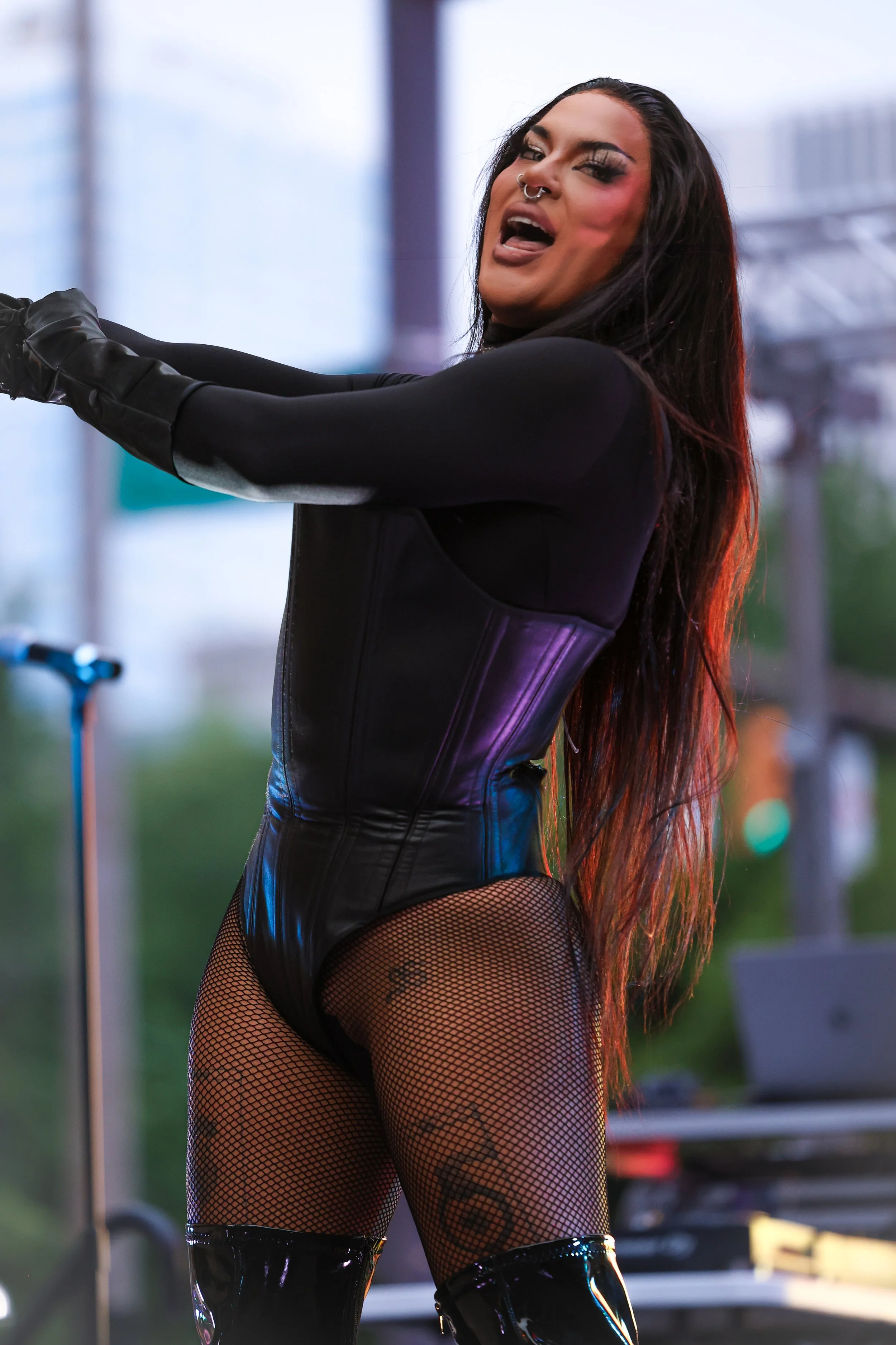 A woman with long dark hair, wearing a black leather outfit, fishnet stockings, and black boots, performing on stage outdoors.