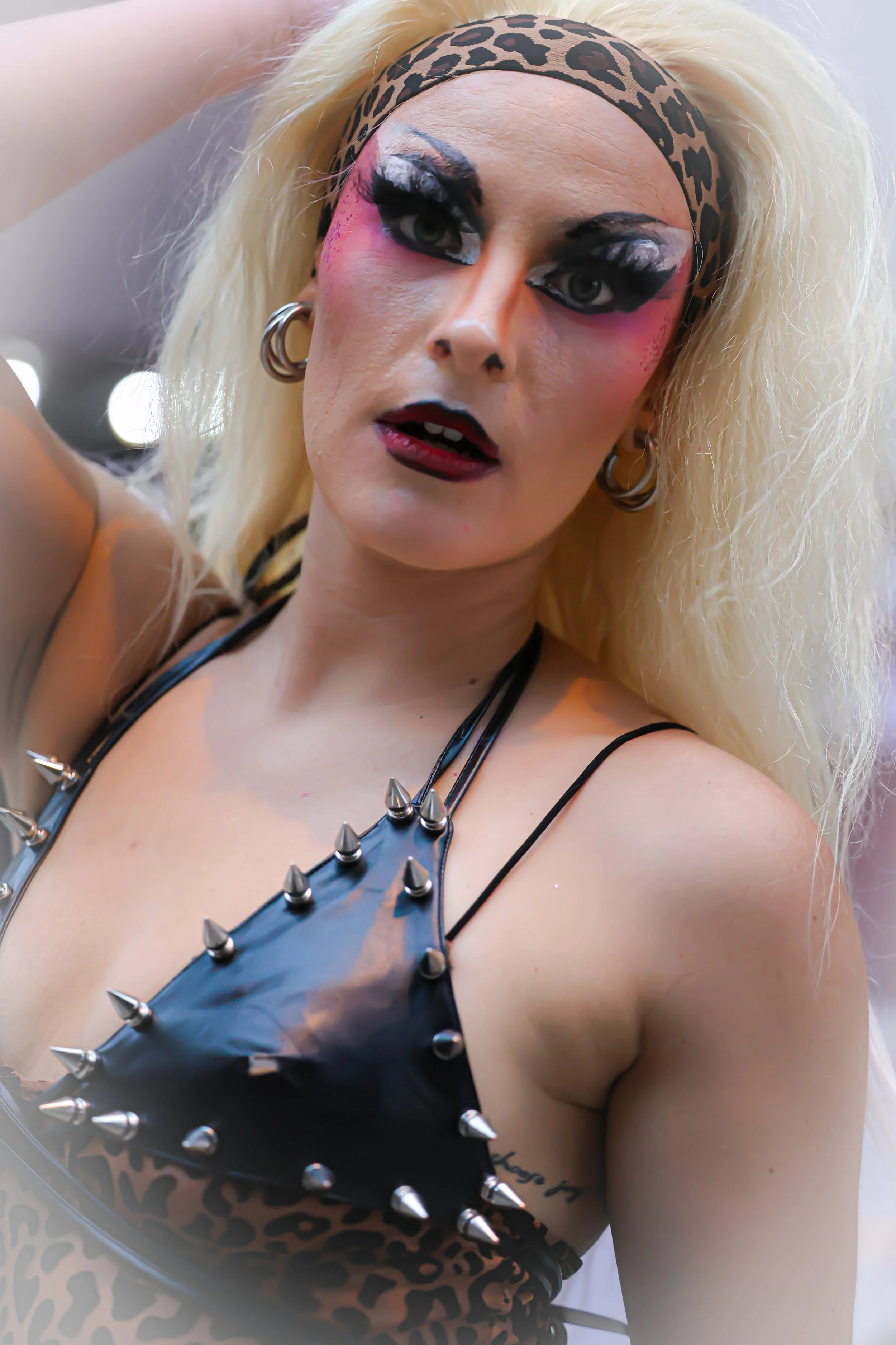 Close-up of a person with platinum blonde, curly hair wearing a leopard print headband, dramatic makeup, and a black leather top with metal spikes.
