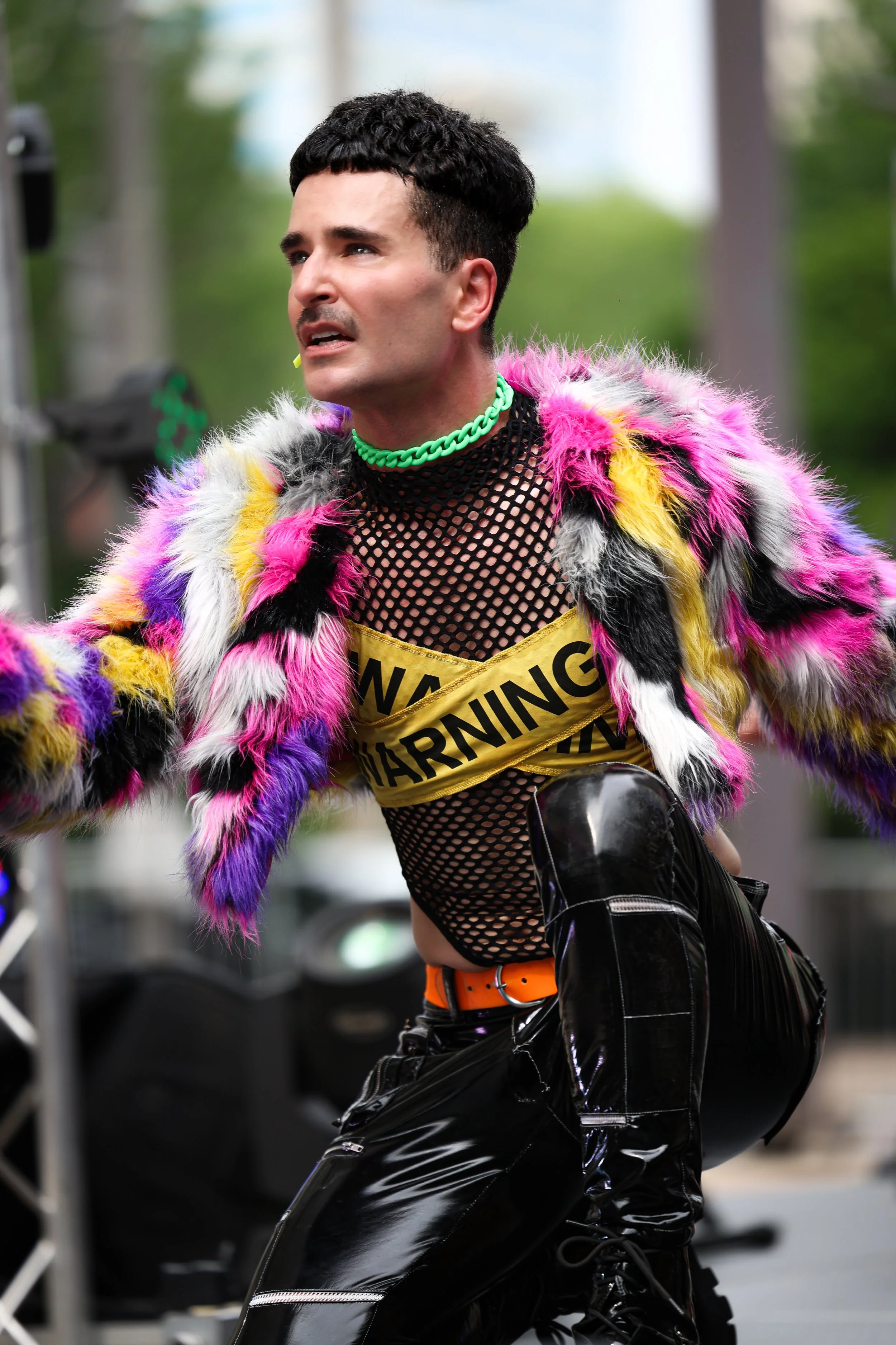 A person is dressed in colorful, flamboyant clothing with a multicolored furry jacket, a yellow 'warning' tape across their chest, and shiny black pants. They are outdoors with a blurred background of green trees.
