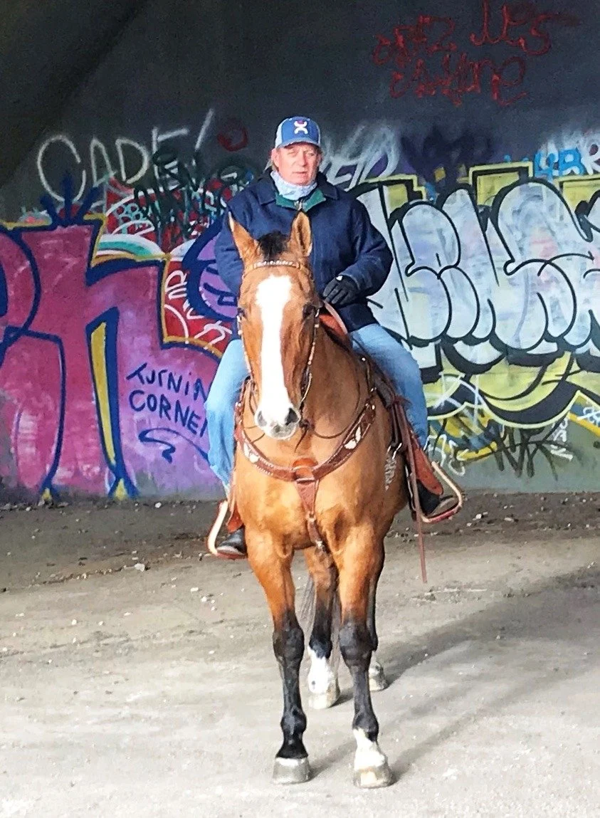 A man wearing a hat and jacket riding a brown horse with a white stripe down its face in front of a graffiti-covered wall.
