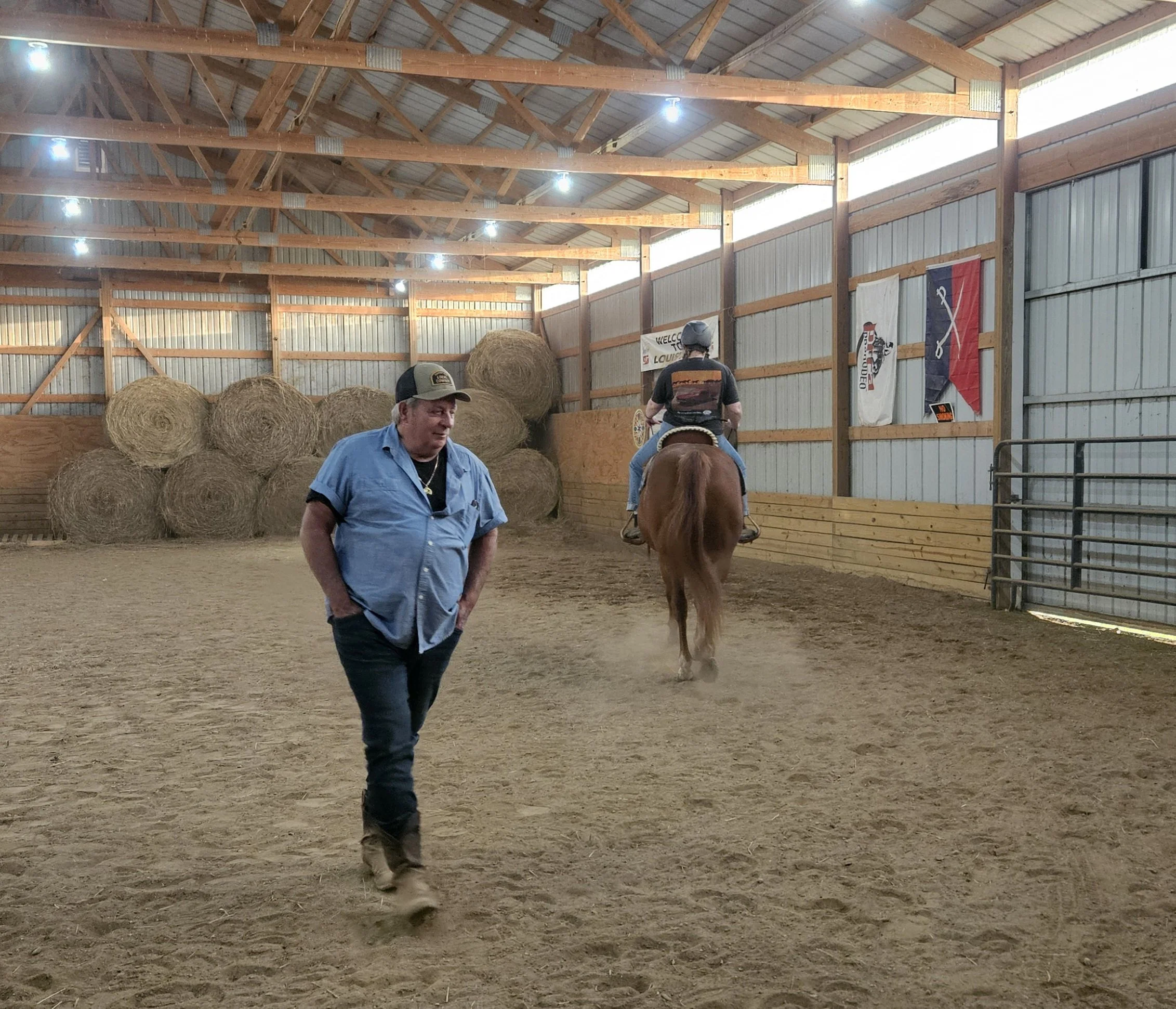 A man walking inside a barn with hay bales in the background, while another person rides a horse.