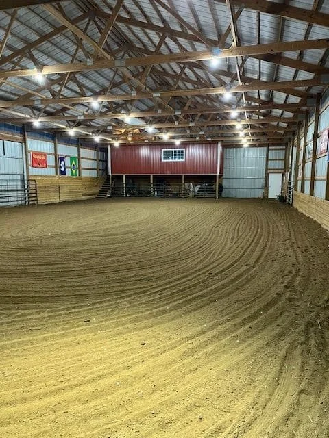 Empty indoor riding arena with a dirt floor, wooden walls, and a metal roof, illuminated by ceiling lights.
