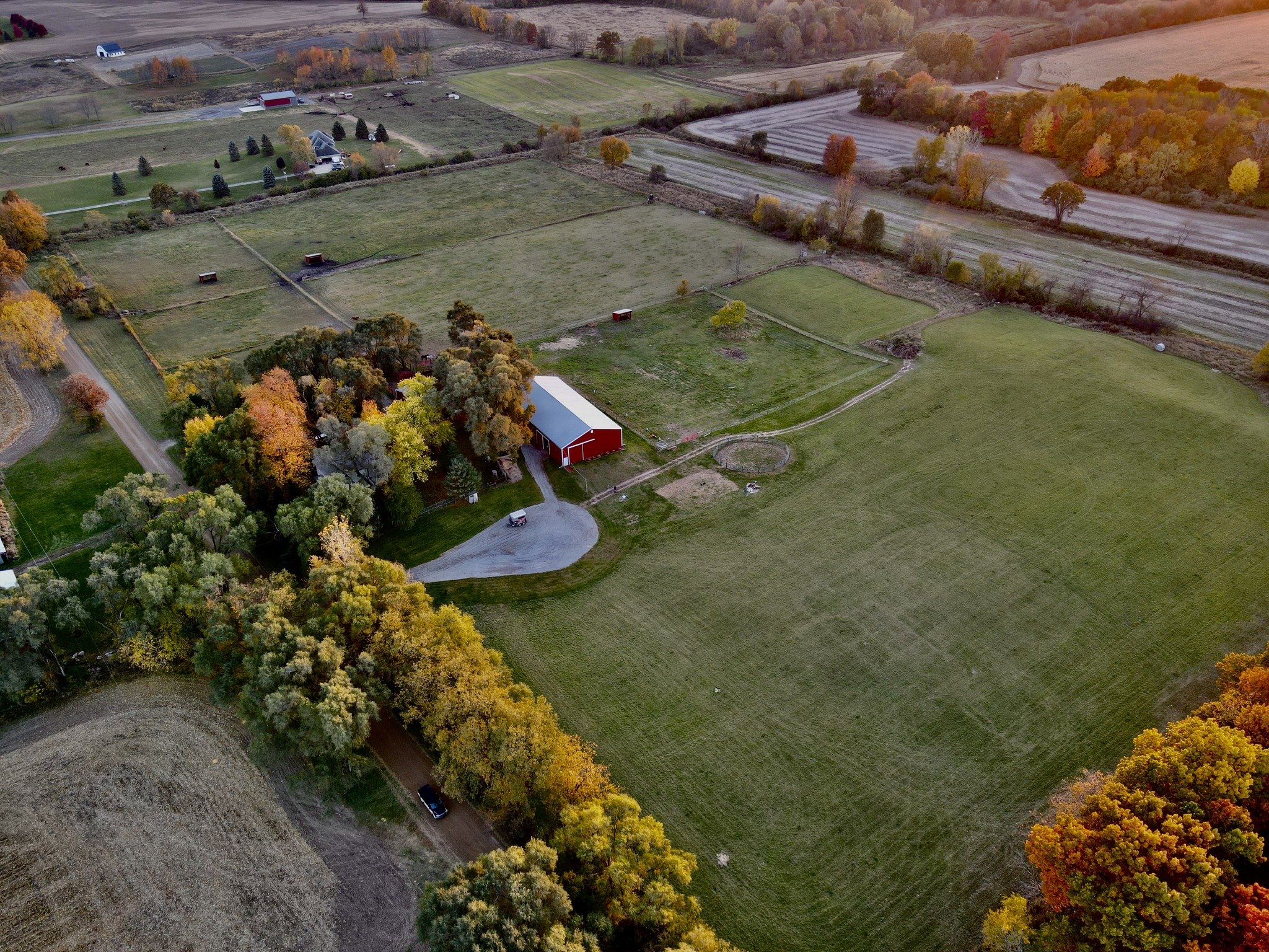 An aerial view of a rural landscape with a red barn, green fields, trees with fall foliage, and farm buildings during sunset.