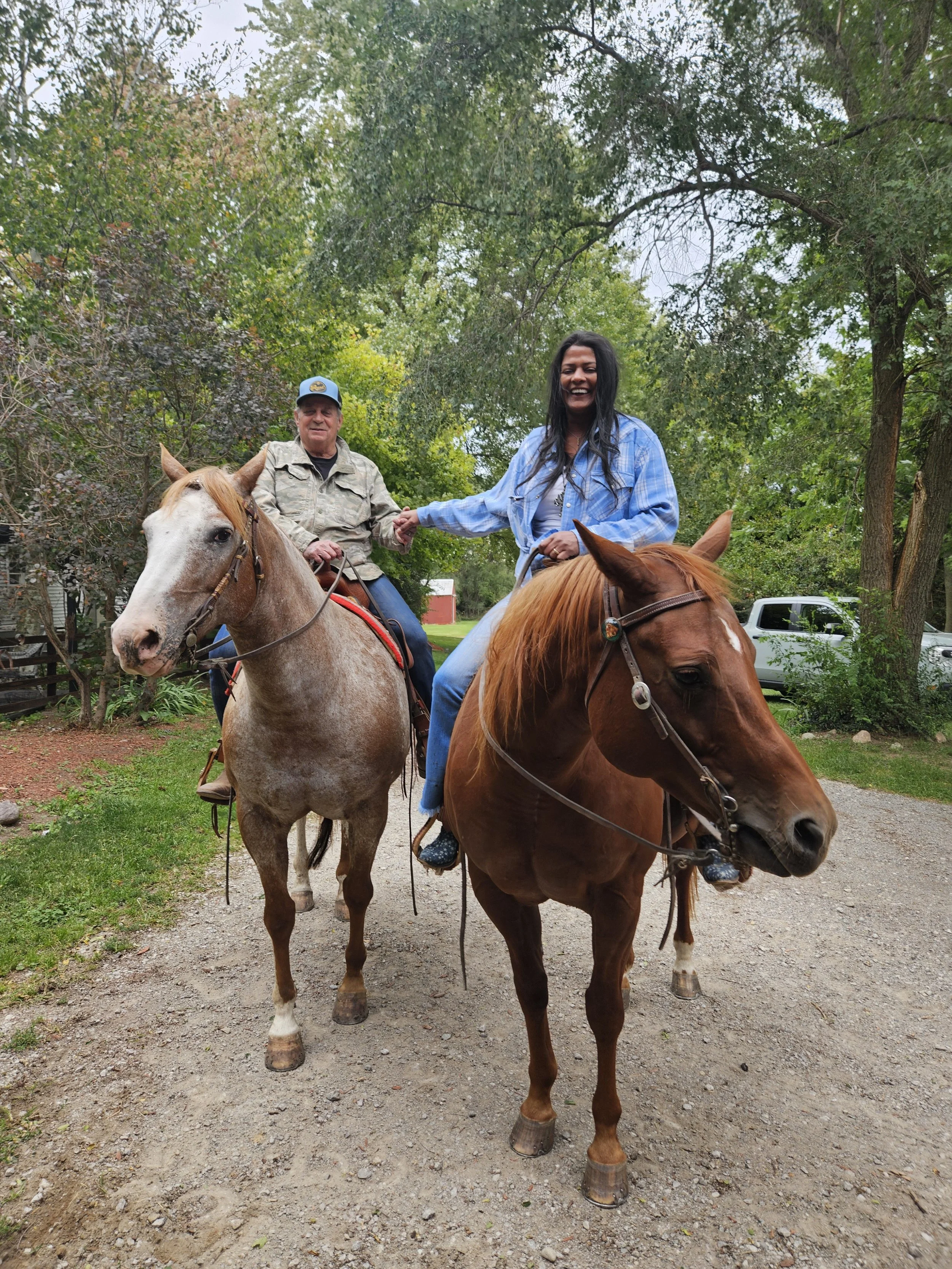Two people riding horses, one man and one woman, outdoors on a dirt path surrounded by trees. The woman is smiling and wearing a blue jacket, while the man is wearing a beige jacket and a cap. They are holding hands and appear to be enjoying a horseback ride.