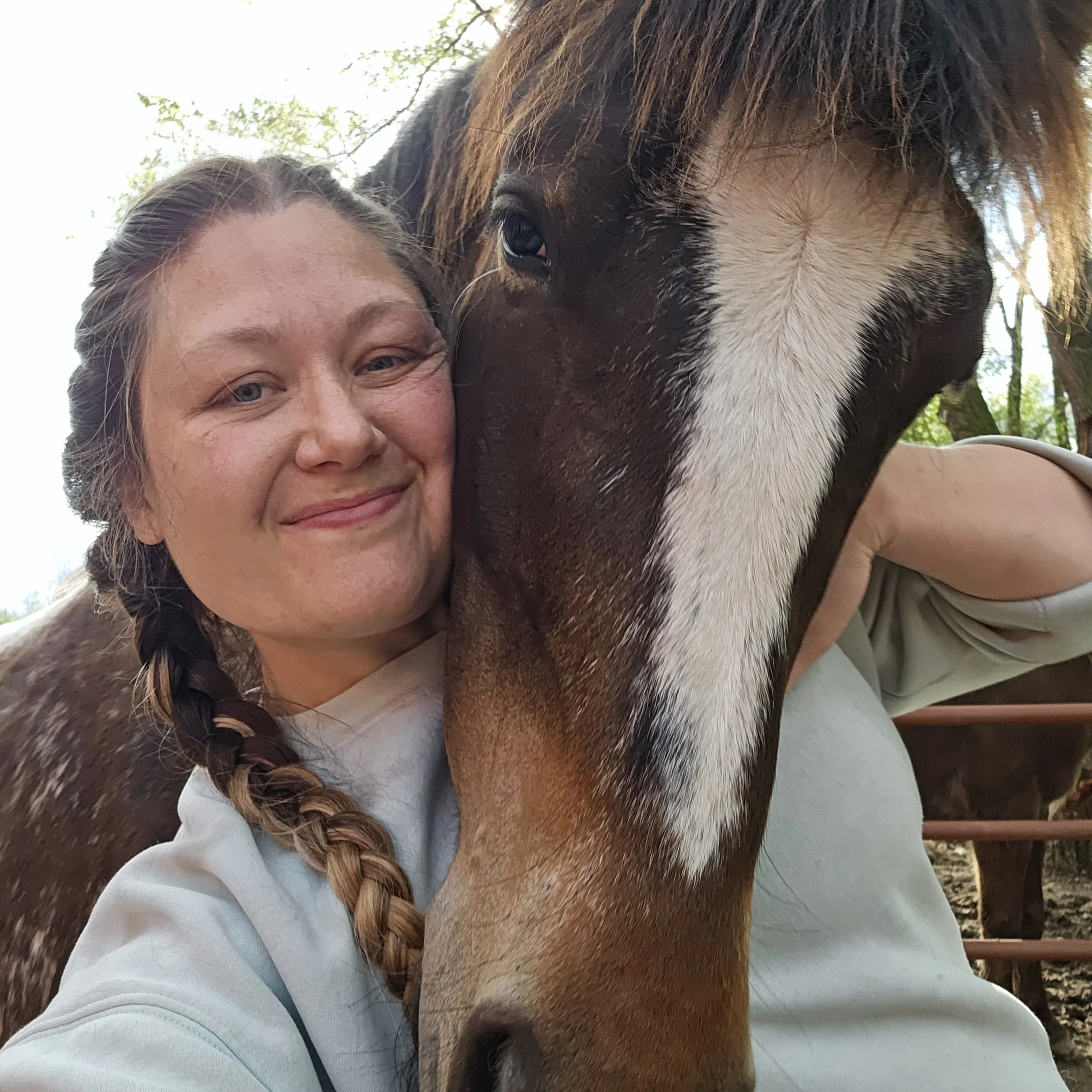 Woman with braided hair smiling and hugging a brown and white horse outdoors.