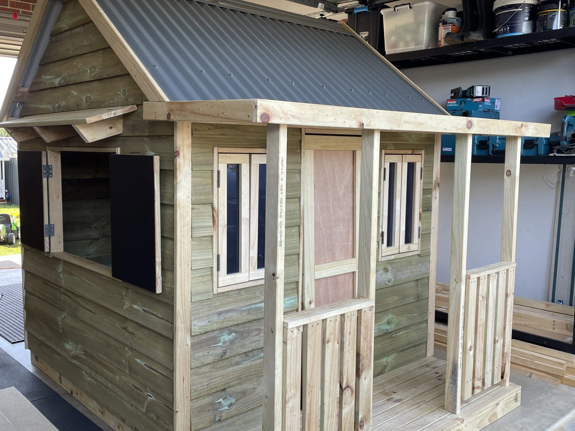 A partially built wooden dollhouse with black window frames, a metal roof, and a small porch area, inside a workshop or garage.