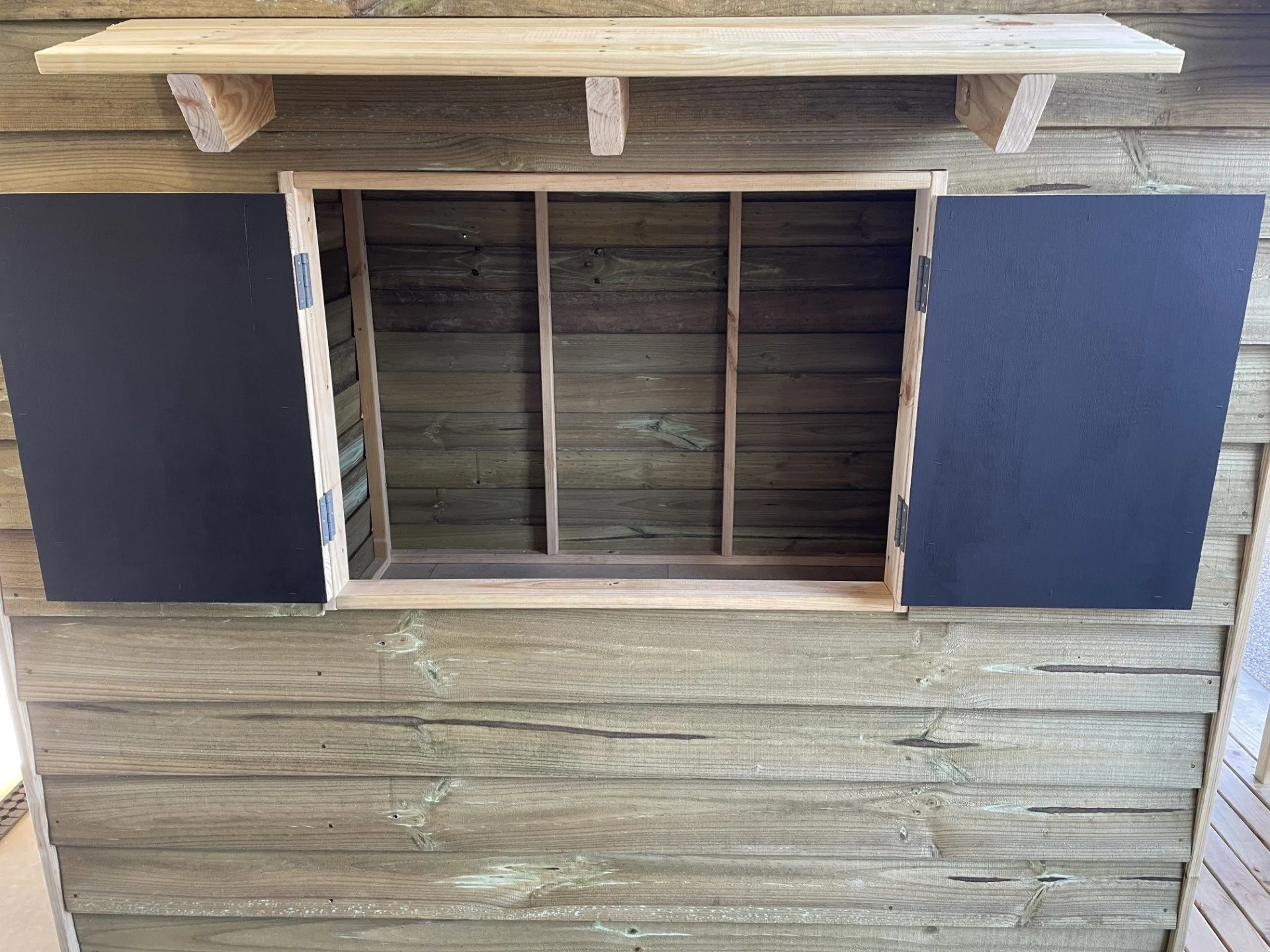 A wooden cubby house with open doors revealing an empty interior, placed against a wooden wall with a horizontal siding pattern.
