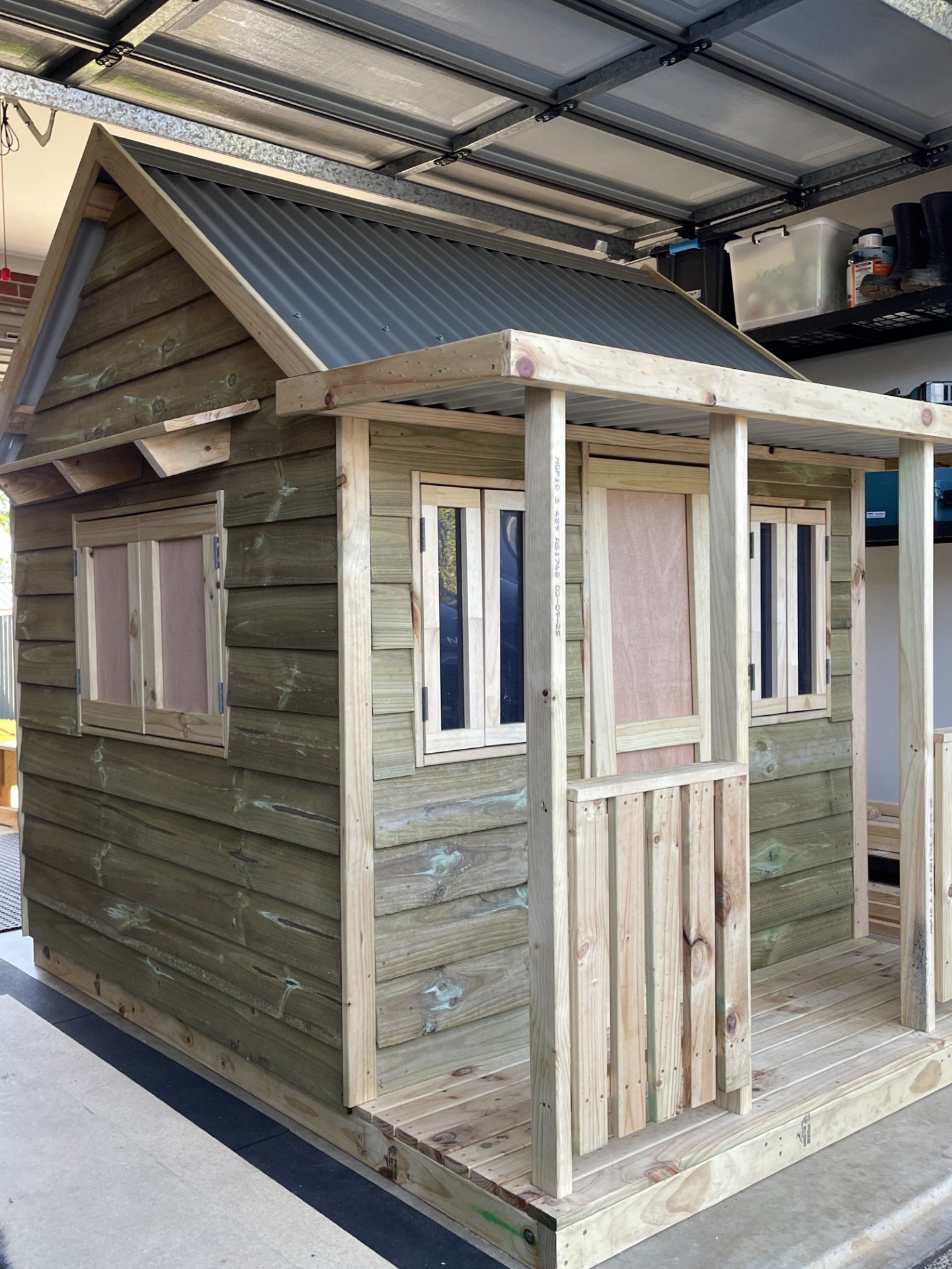Unfinished wooden playhouse with a metal roof, small porch, and windows, situated inside a garage.