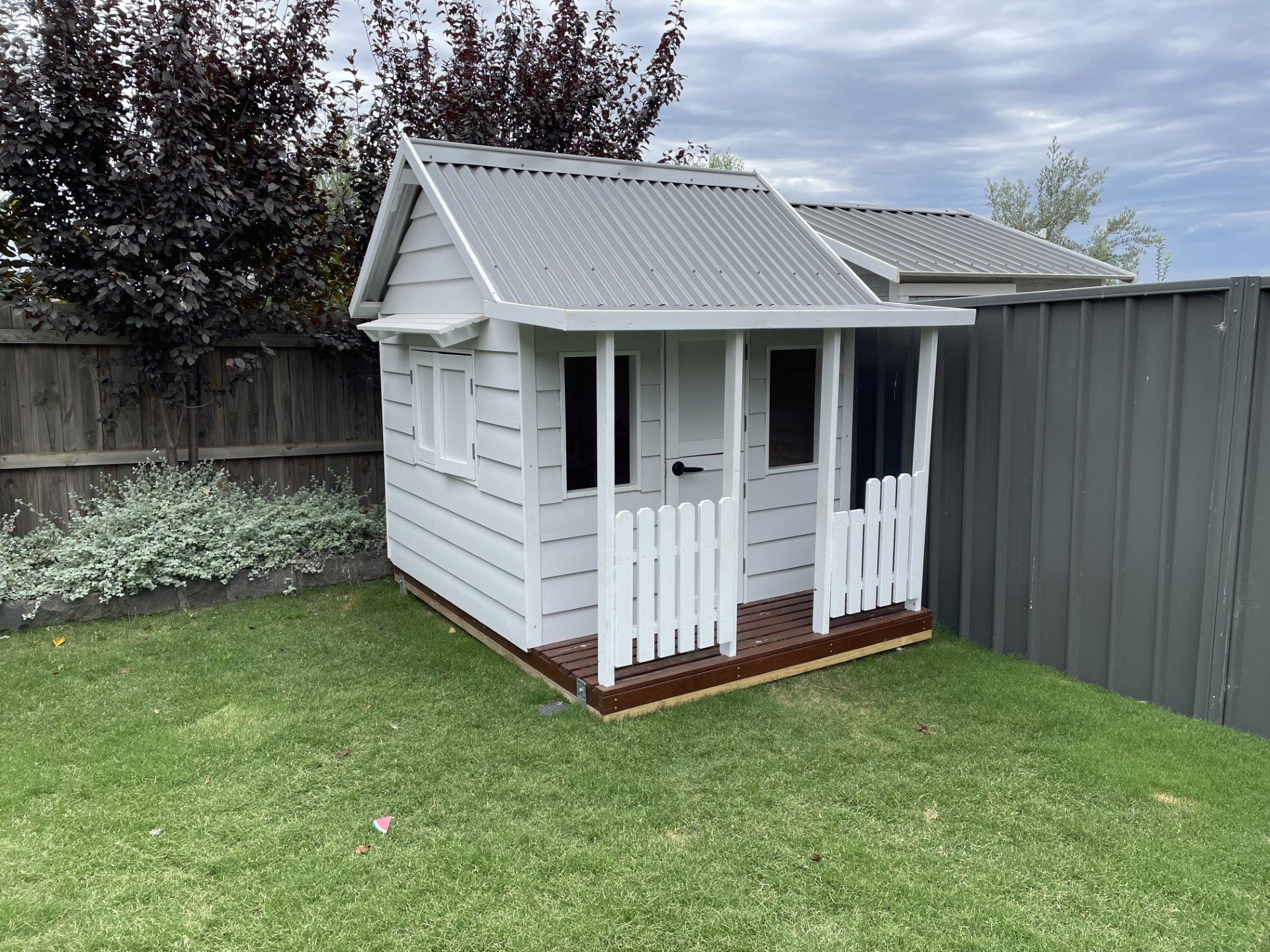 A small white wooden playhouse with a mini porch, situated on a grassy backyard, fencing, and bushes, under a cloudy sky.