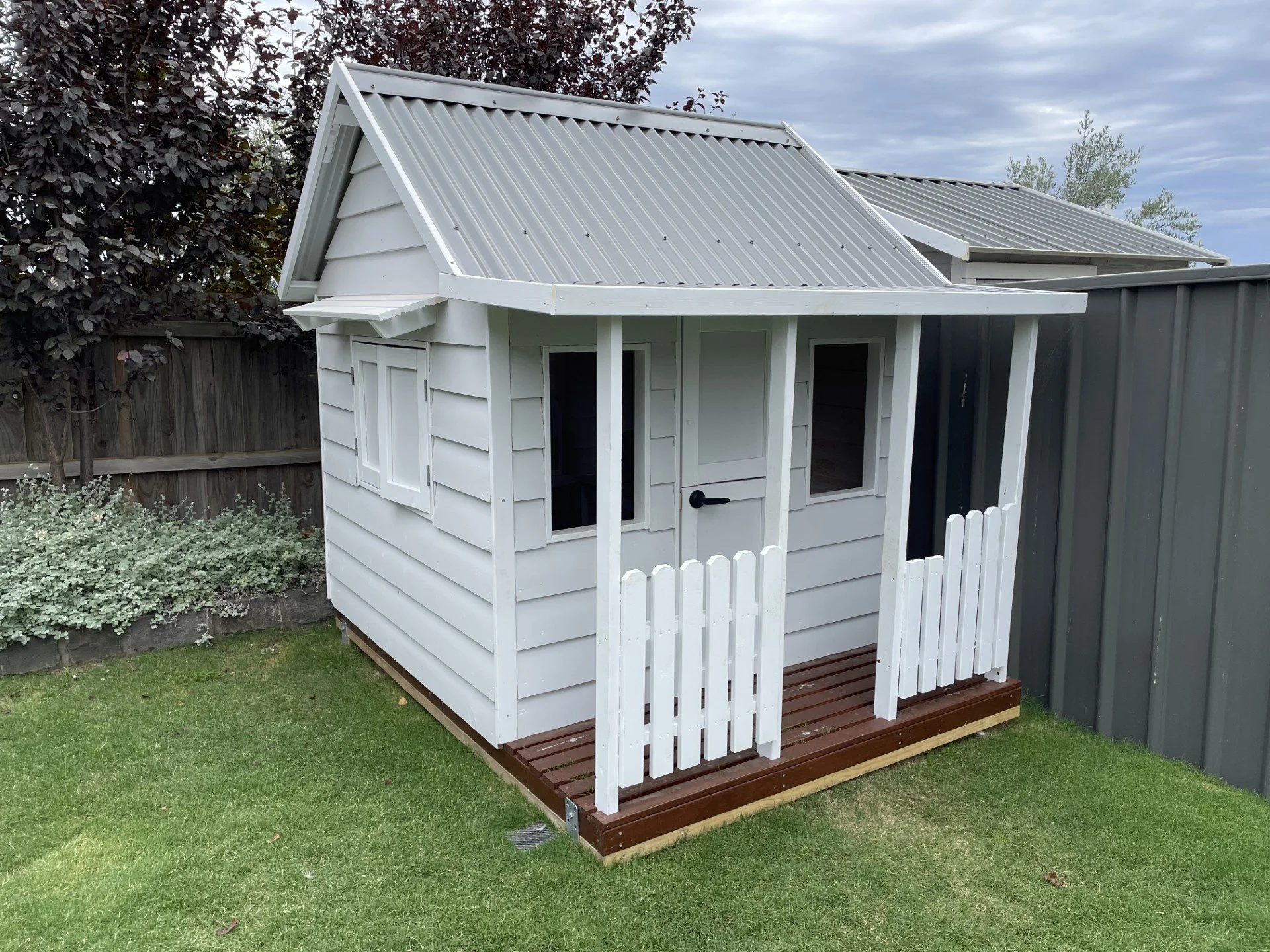 Small white wooden playhouse with a deck, fenced backyard, trees, cloudy sky, and metal roofing.
