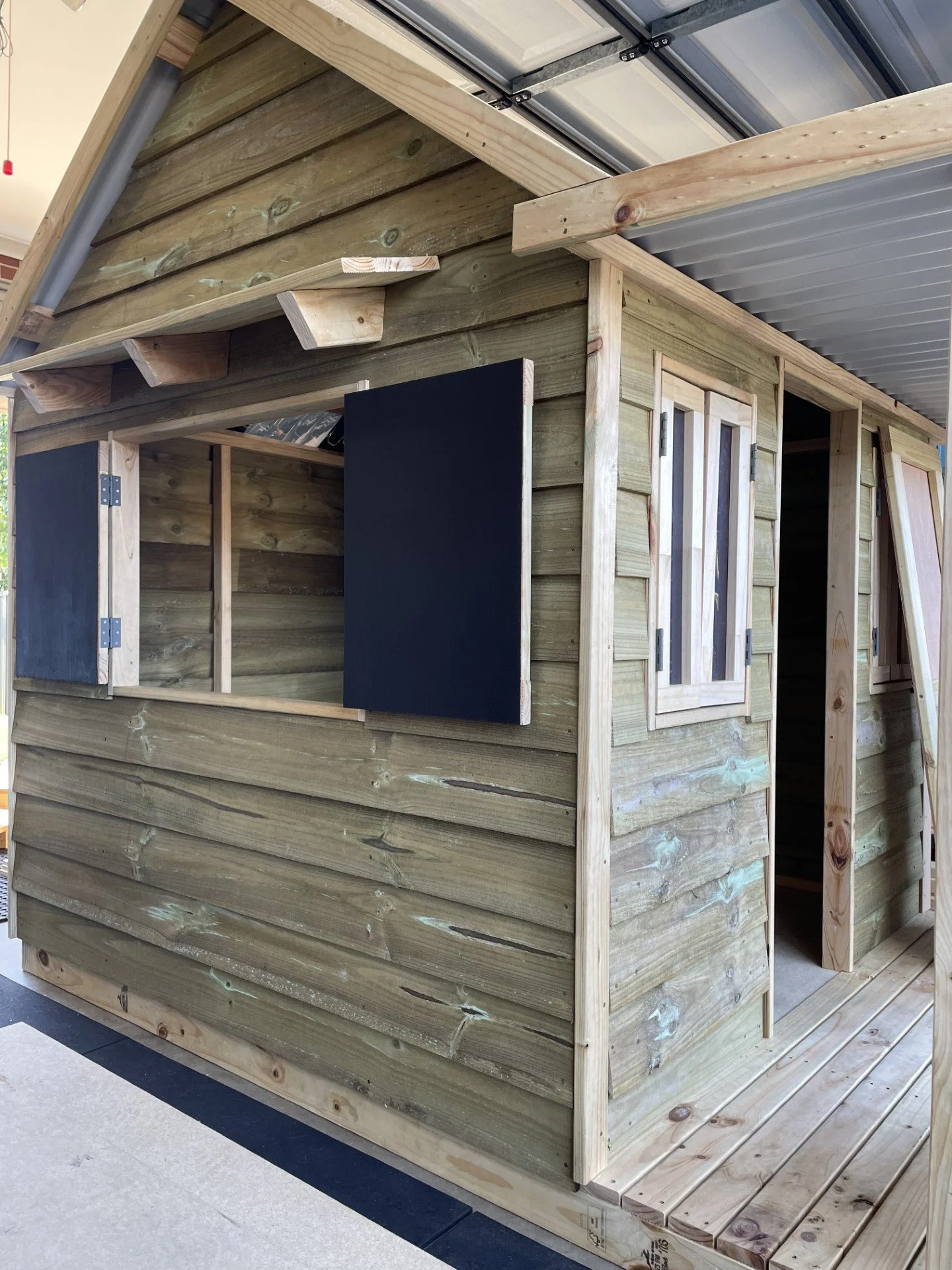 A wooden playhouse under construction with partial siding, windows, and a door frame, located indoors on a wooden floor