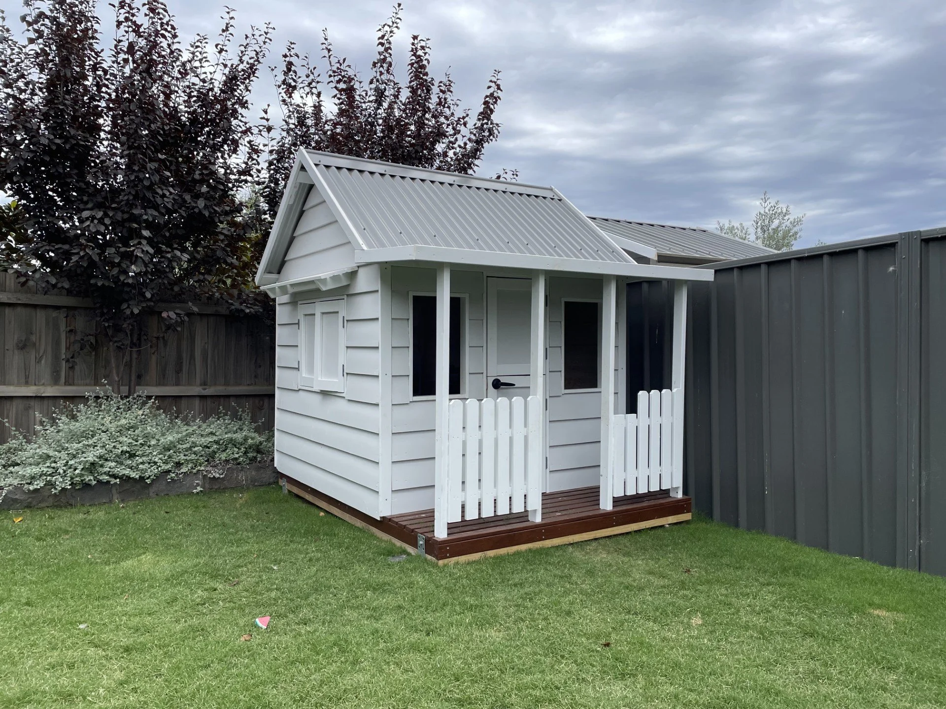 Small white wooden playhouse with a porch, located in a yard with green grass and a flower bed with shrubs. Dark fence and trees in the background under a cloudy sky.