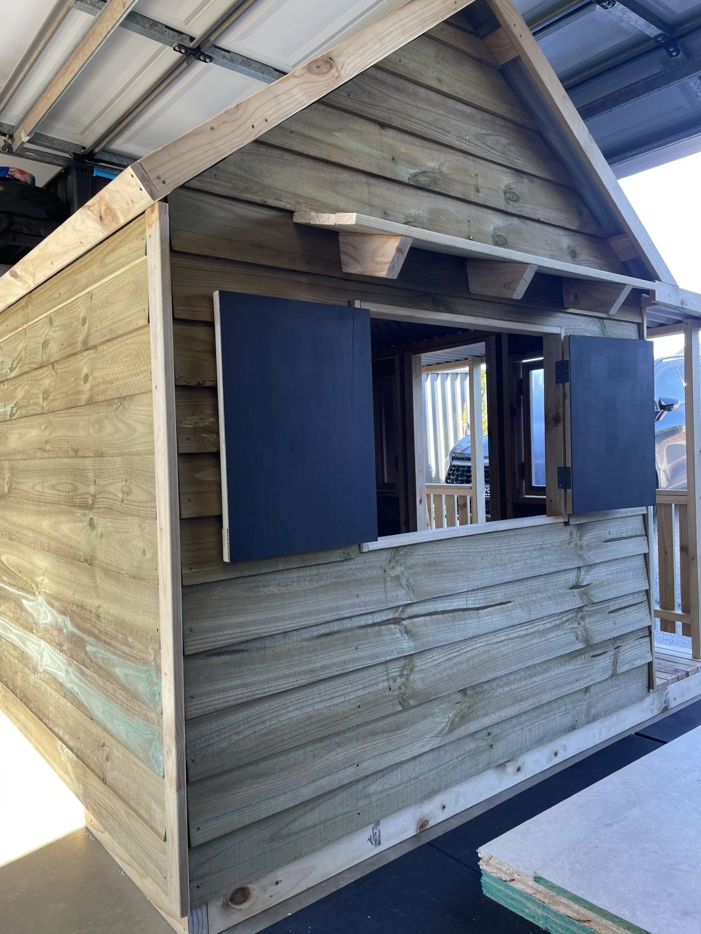 The image shows a small wooden structure under construction, with the exterior walls made of horizontal wooden planks and a partially open window with dark shutters.
