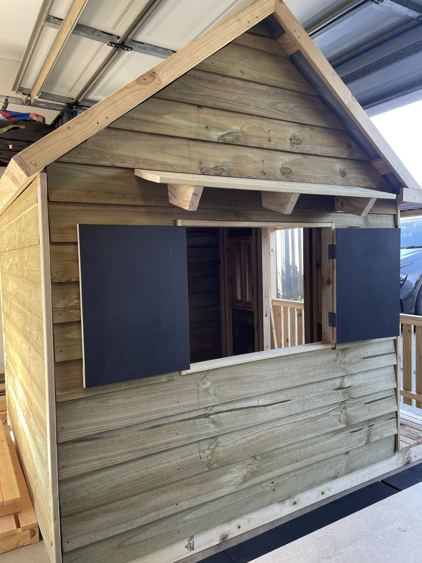 A small wooden playhouse under construction, featuring a pitched roof with exposed wooden beams, an open doorway, and a window with black shutters. The house is built with horizontal wooden planks.
