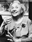 An elderly woman smiling and holding a coffee cup, wearing a blouse with a flower pin.