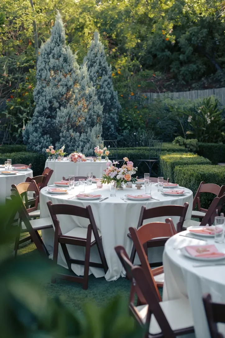 Round reception tables set with white linens and pink florals among the garden hedges.