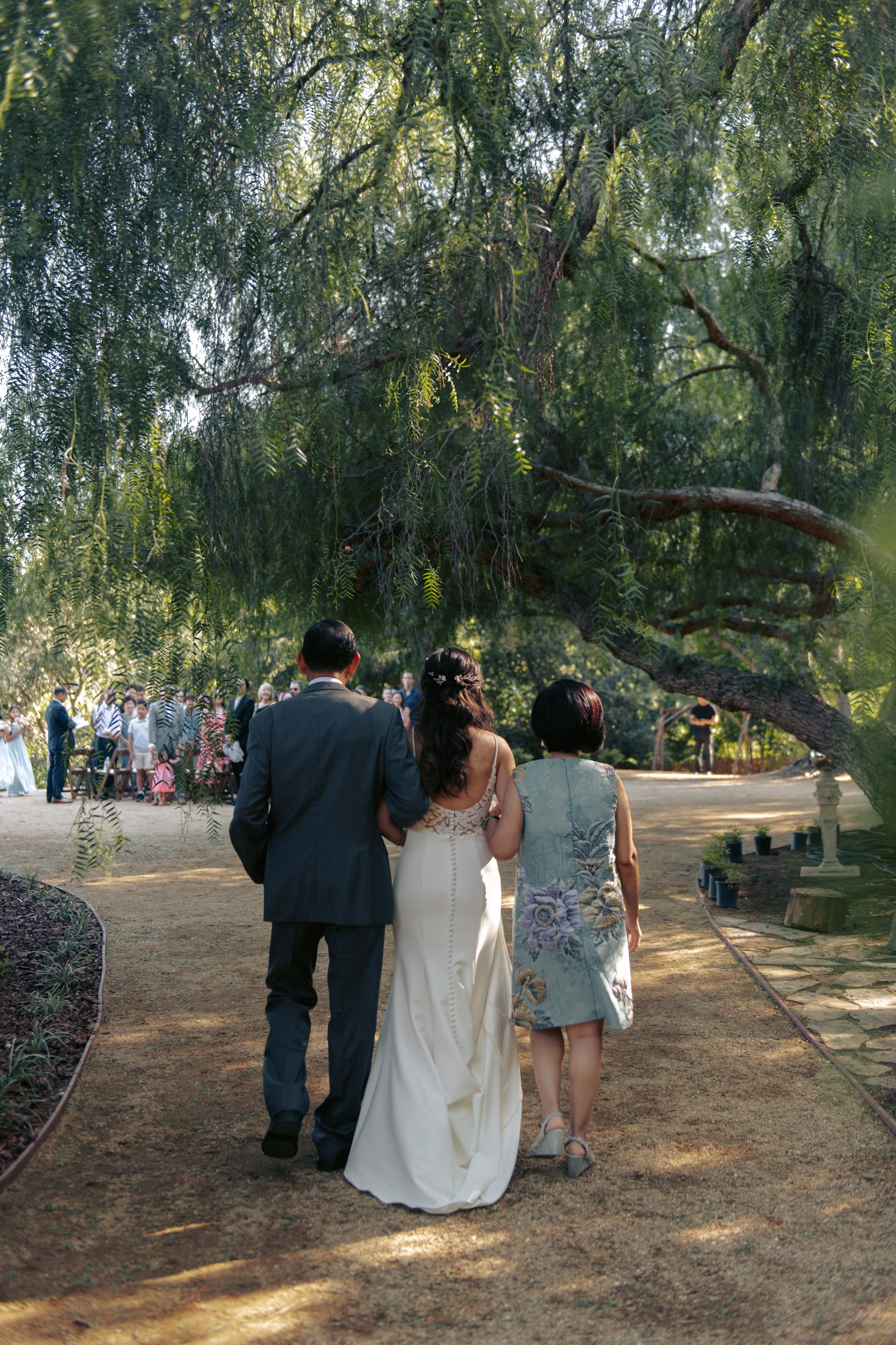 Newlyweds walking a garden path with family, past guests seated beneath a willow tree.