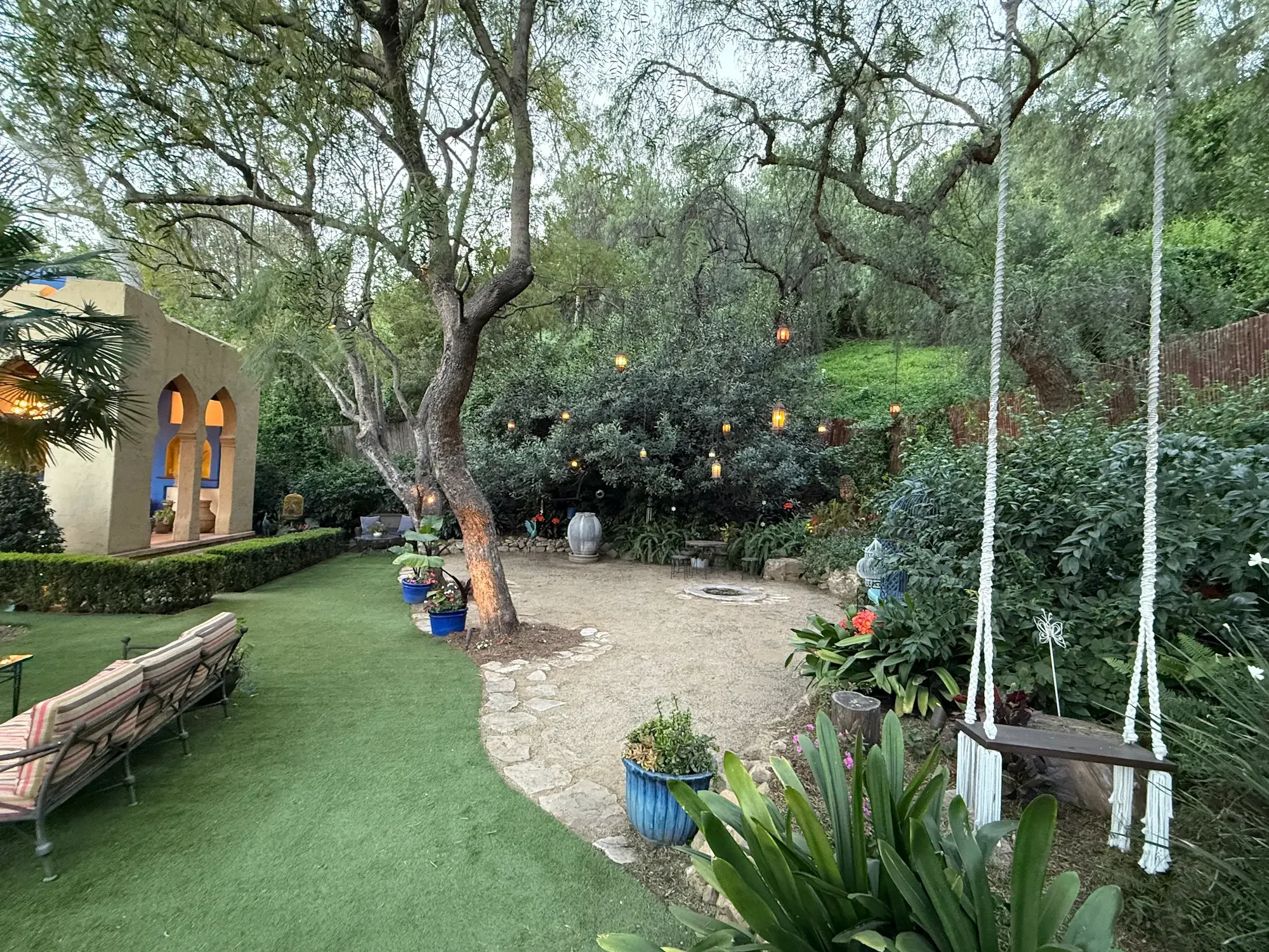 Garden setting with a macramé swing, fire-pit lounge, and Moroccan lanterns hanging beneath mature trees.