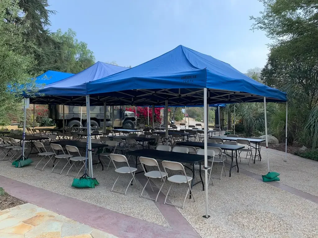 Tables and chairs beneath blue canopies in the motor court at XOHH Events, set for a catered lunch serving over 120 crew members, with cooking staged outside the gate at this Los Angeles filming location.
