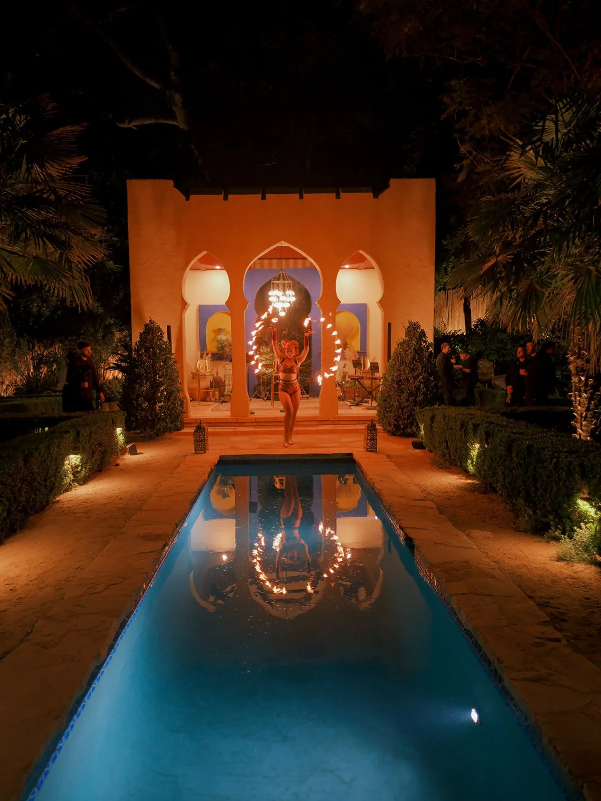 Moroccan pavilion glowing at night, reflected in the pool, a figure walking the lit path