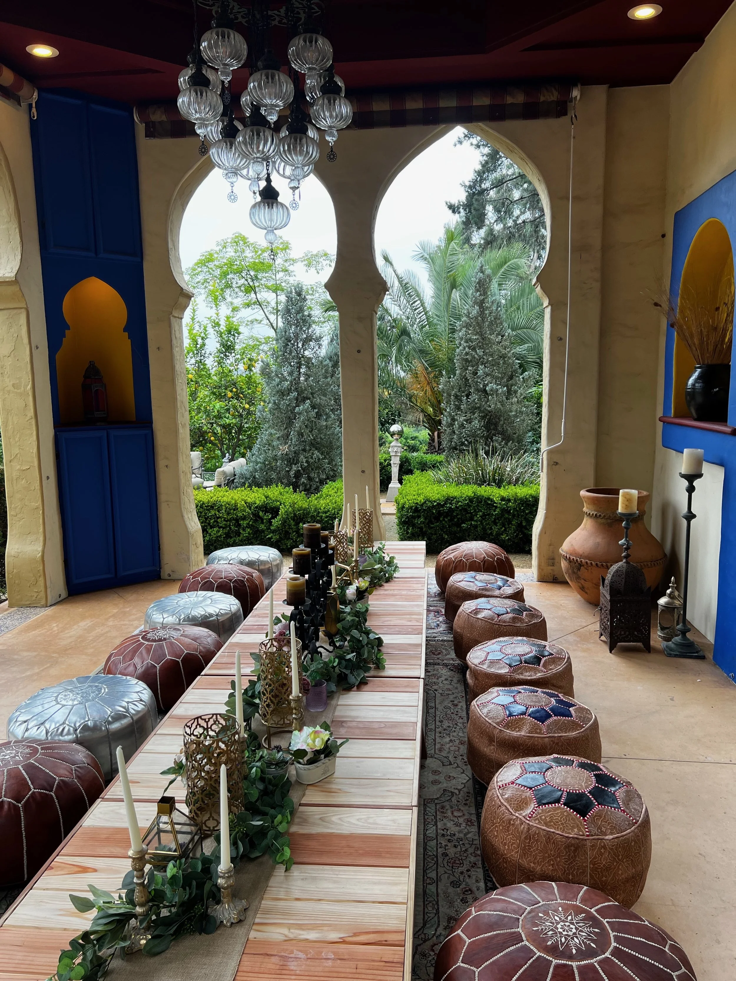 Low Moroccan banquet table with candles and trailing greenery flanked by leather poufs, beneath a glass chandelier and keyhole arches opening to the gardens.