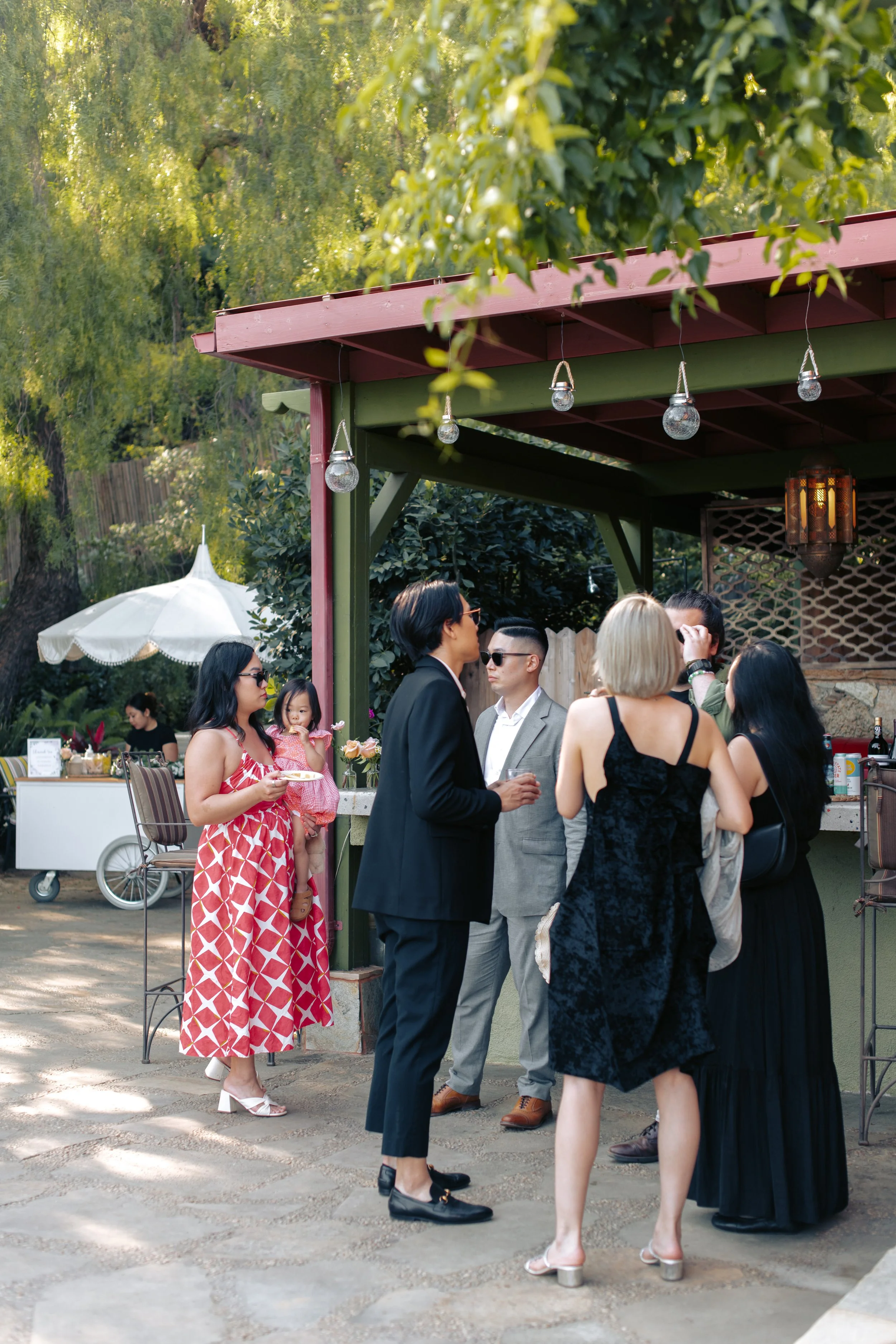 Guests gathering under a lantern-hung garden pergola near a dessert cart and white umbrella.