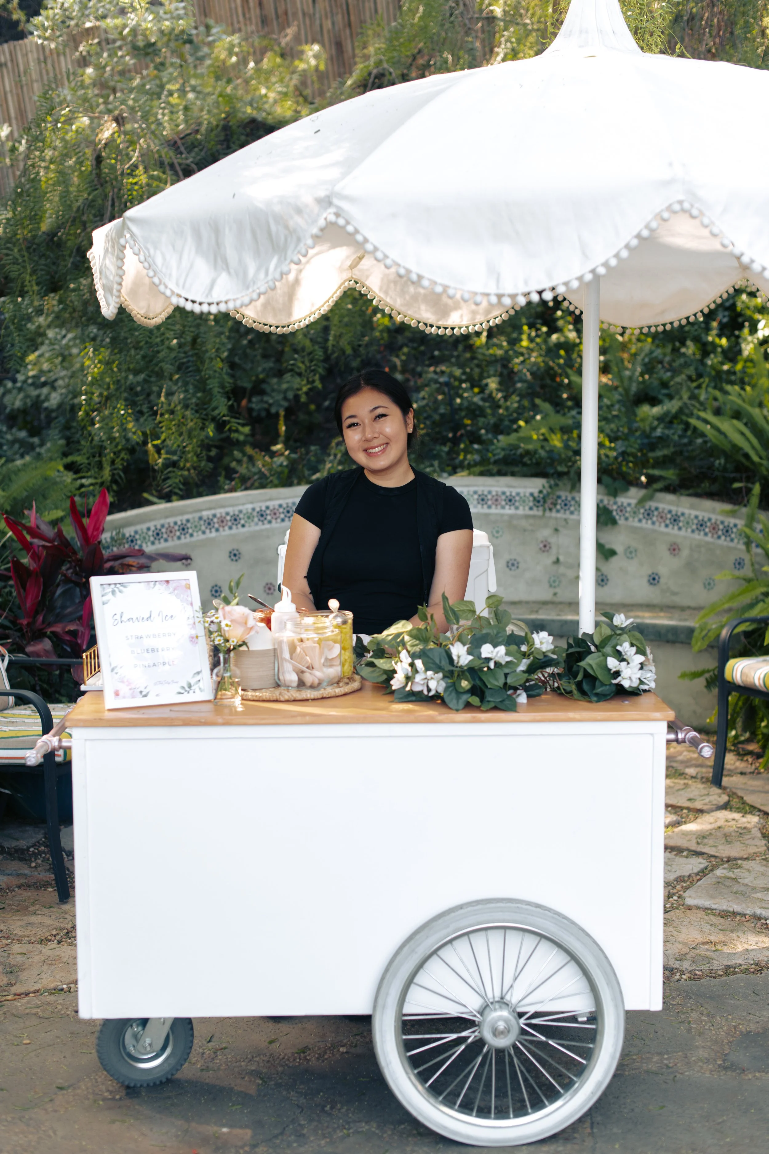 Attendant behind a white vintage cart with a scalloped umbrella and floral trim beside the tiled fountain.