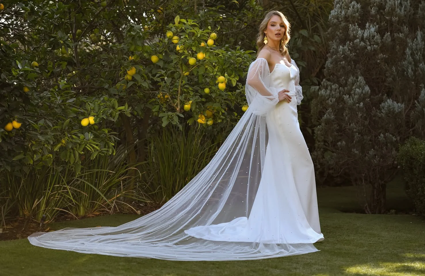 Bride in a sheer-sleeved gown with a long veil beside the lemon tree.