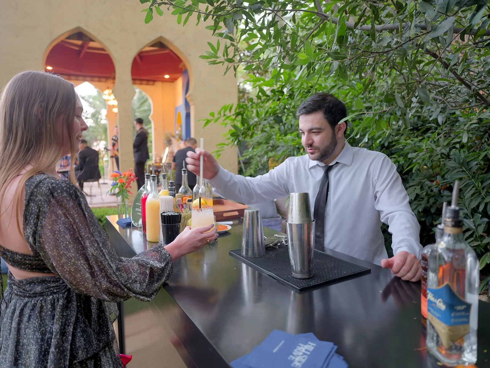 Bartender mixing cocktails at an outdoor bar as a guest reaches for a drink