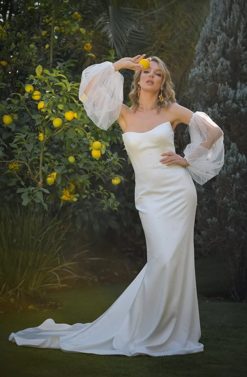 Bride in a sheer-sleeved gown holding a lemon, posing beside a fruiting lemon tree in the garden.