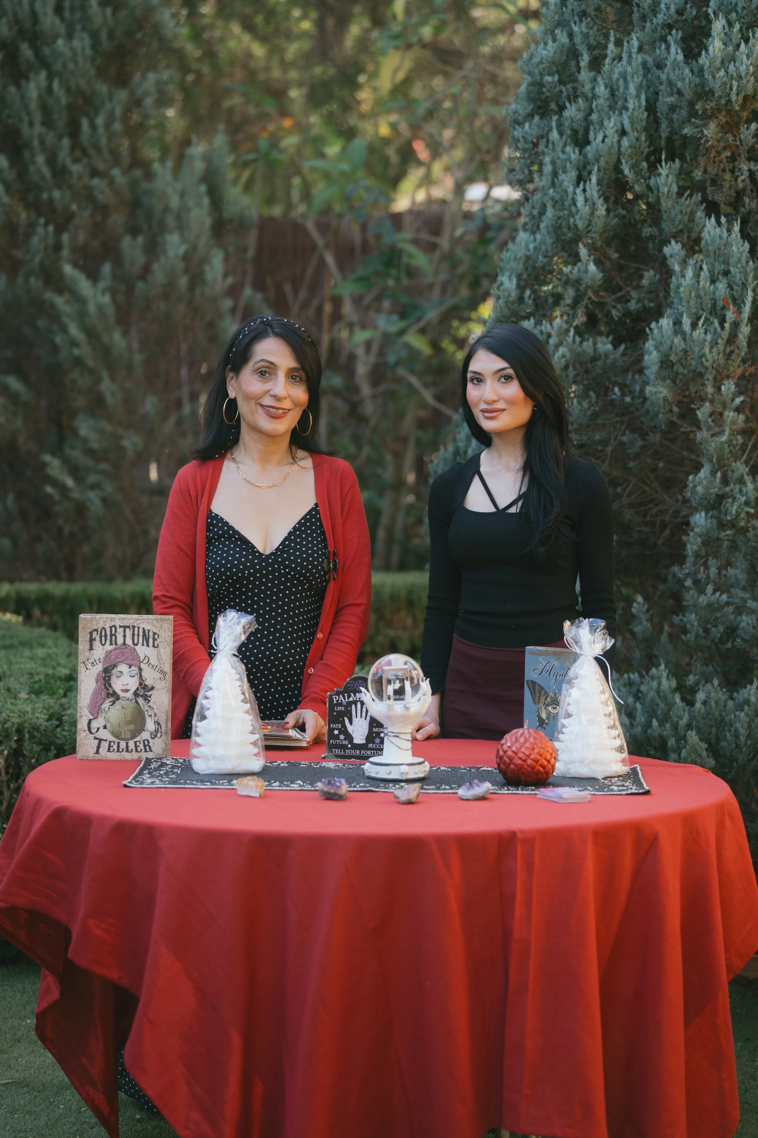Two hosts behind a red-draped "Fortune Teller" table with a crystal ball and tarot cards in the garden.