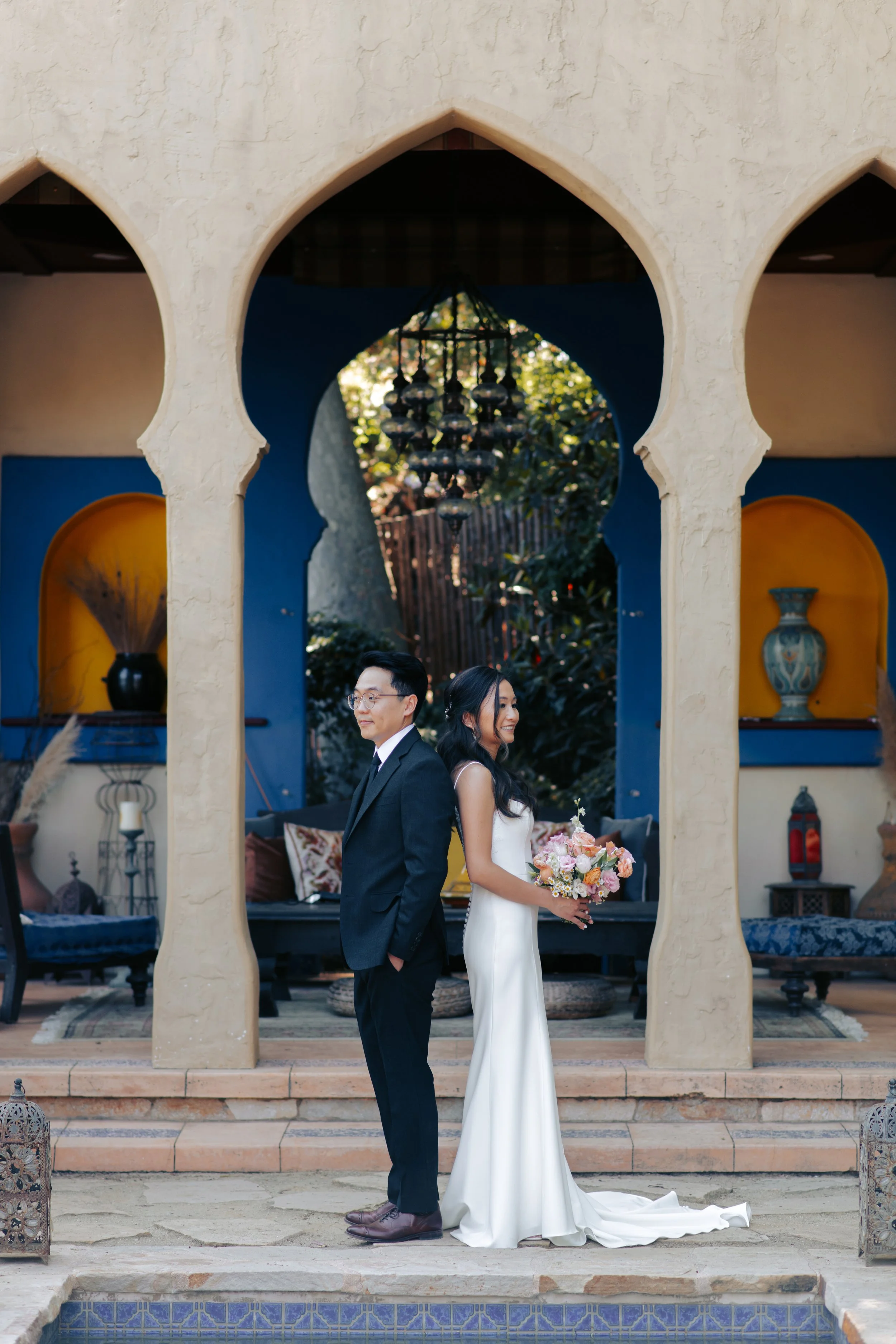 Bride and groom standing back-to-back beneath a Moroccan keyhole archway with cobalt walls and a hanging chandelier, the bride holding her bouquet.