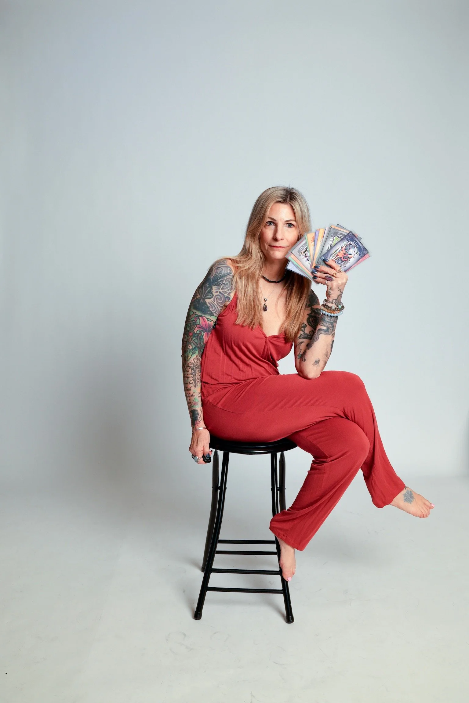 A woman with long blonde hair, tattoos on her arms and legs, wearing a red outfit, sitting on a black stool, holding a fan of tarot cards, against a plain light gray background.