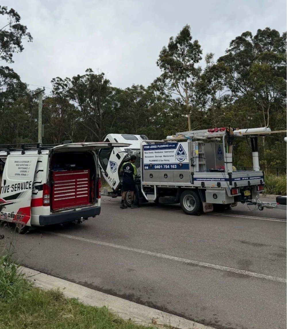Two service vehicles, one with its rear open and a worker in uniform, on a road with traffic and trees in the background.